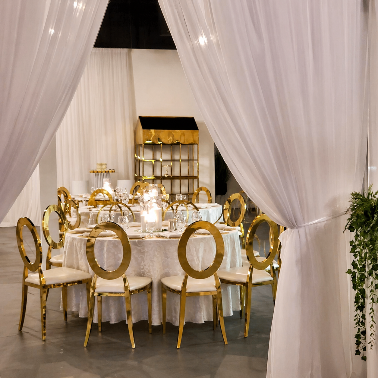 Elegant banquet table setup with gold-rimmed chairs, white tablecloth, candles, and glassware, seen through white draped curtains at a formal event.
