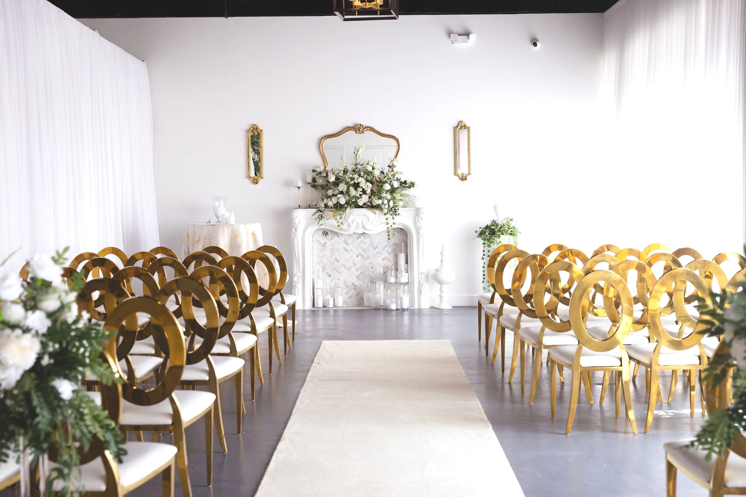 Elegant wedding ceremony setup with golden chairs facing a white fireplace adorned with white flowers and greenery, candles, and gold-framed mirrors on a white wall.