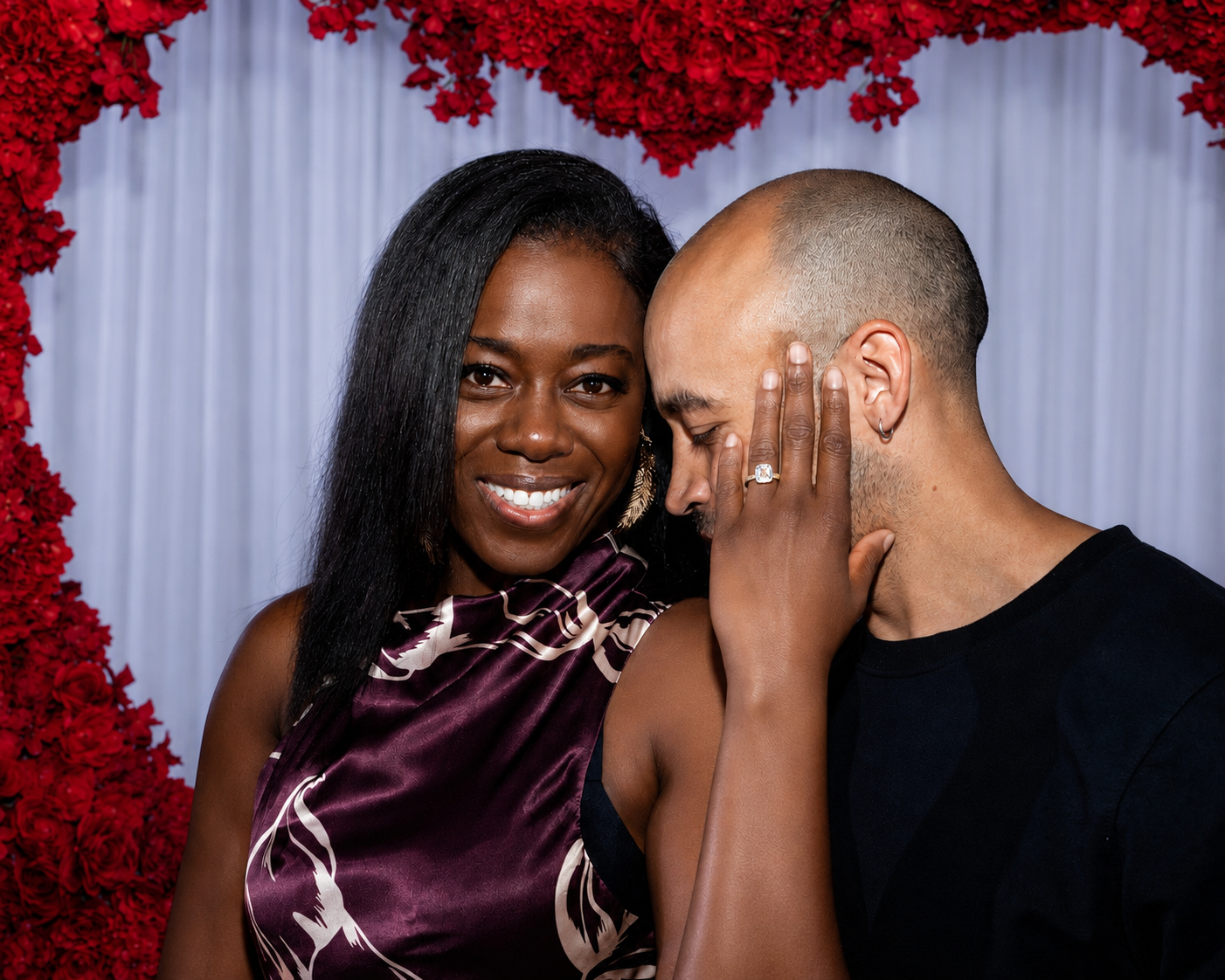 A woman and a man are embracing at a wedding or celebration, with the woman smiling and the man touching his face. The woman has long black hair and is wearing a purple satin dress, while the man has a shaved head, a nose ring, and is wearing a black shirt. They are standing in front of a background decorated with red flowers and white curtains.