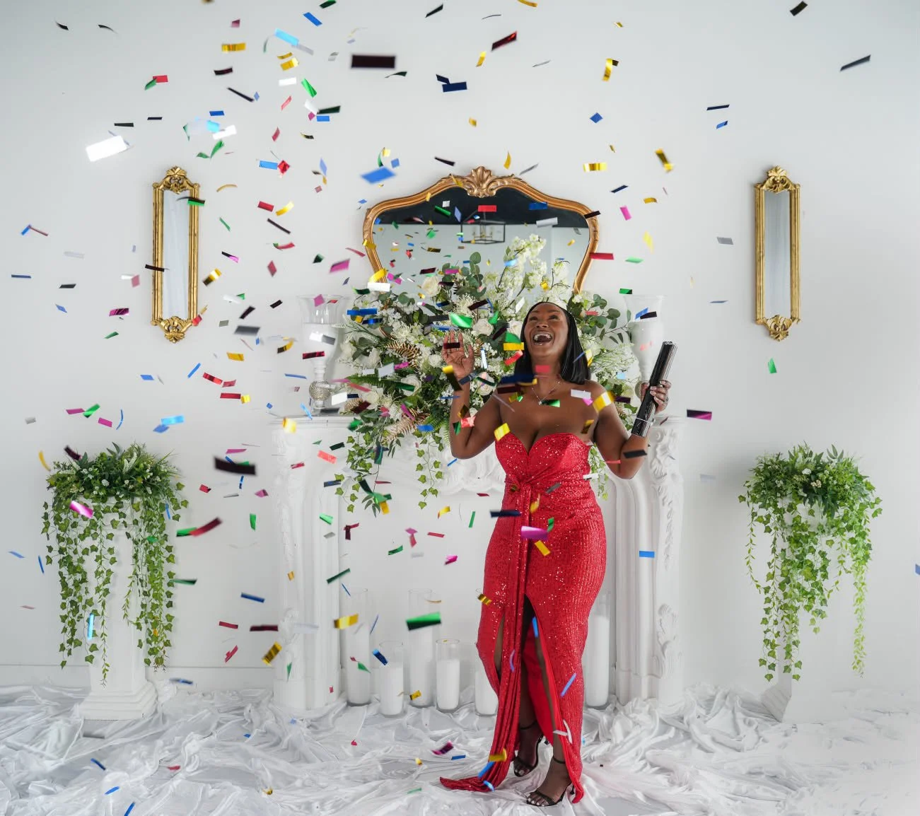 A woman in a red sequined dress celebrating with confetti in a decorated room with green plants and a mirror.