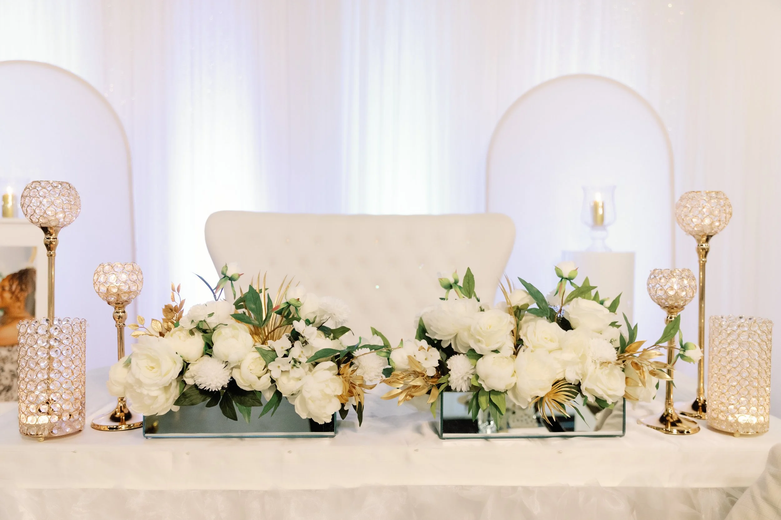 Elegant wedding centerpiece featuring white roses, greenery, and gold accents, with gold and crystal candle holders on a white tablecloth.