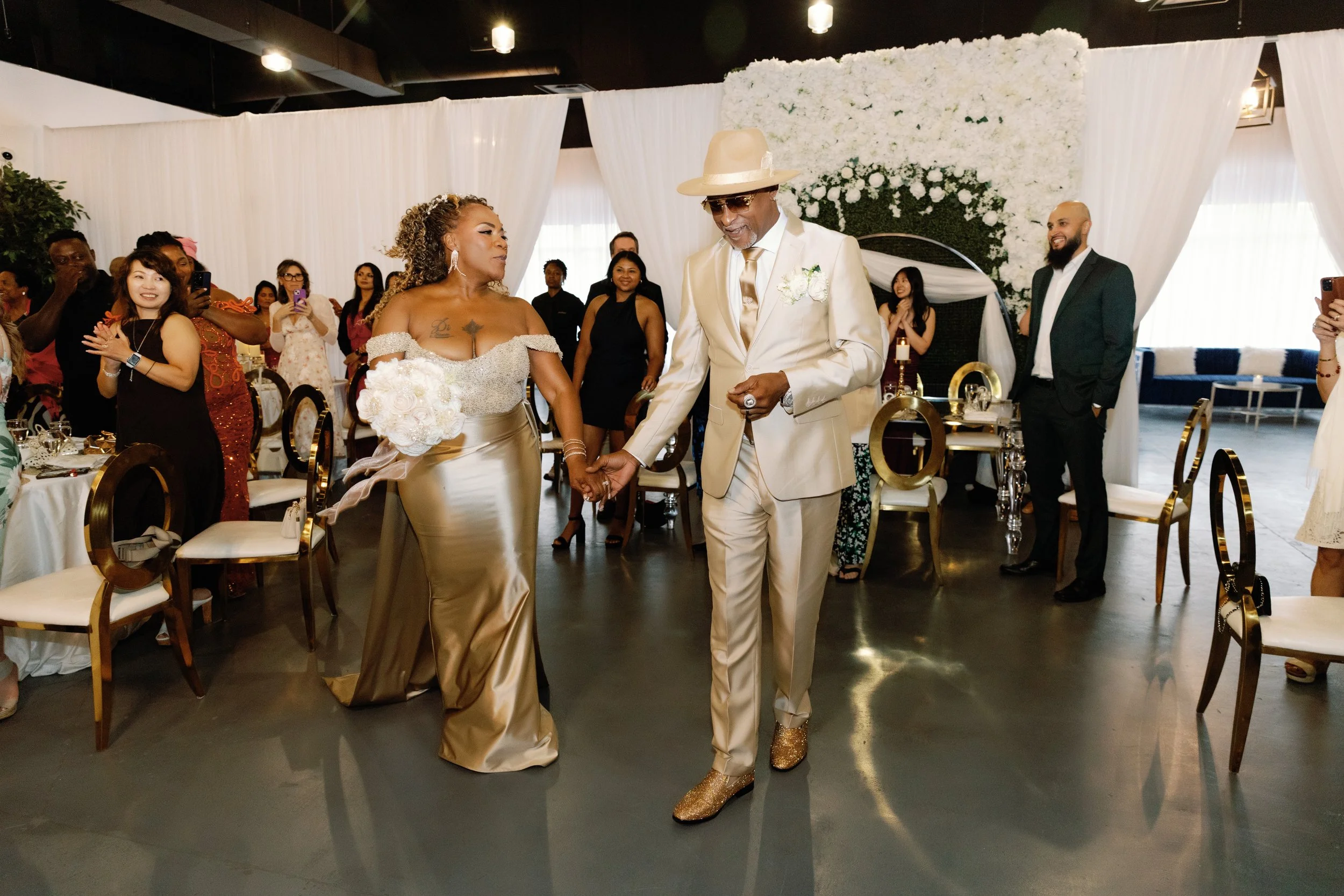 A bride and groom holding hands during their wedding ceremony surrounded by friends and family, with a floral backdrop.