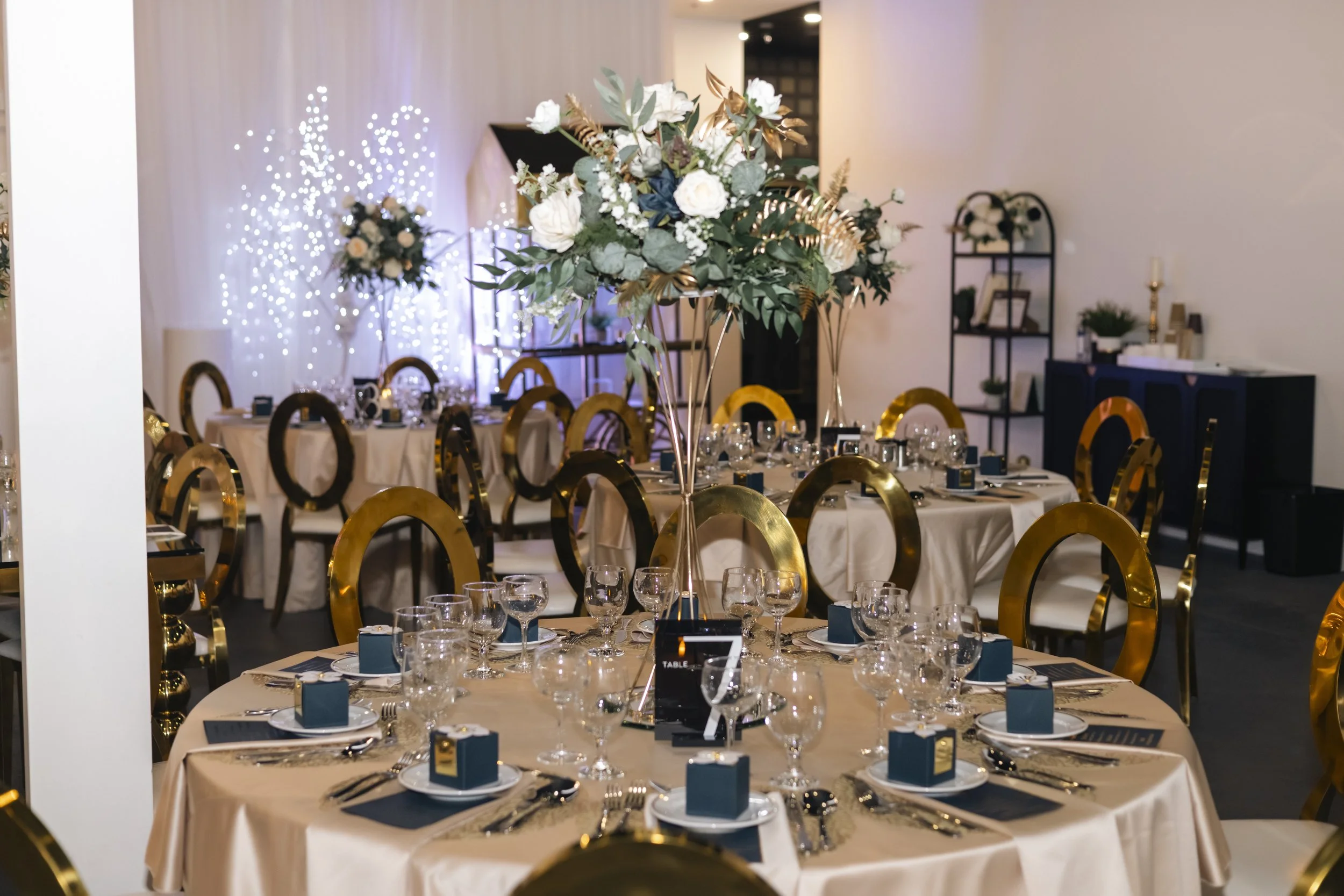 Elegant banquet hall decorated with floral centerpieces, set tables with glassware and napkins, and soft lighting with blue accents and string lights.