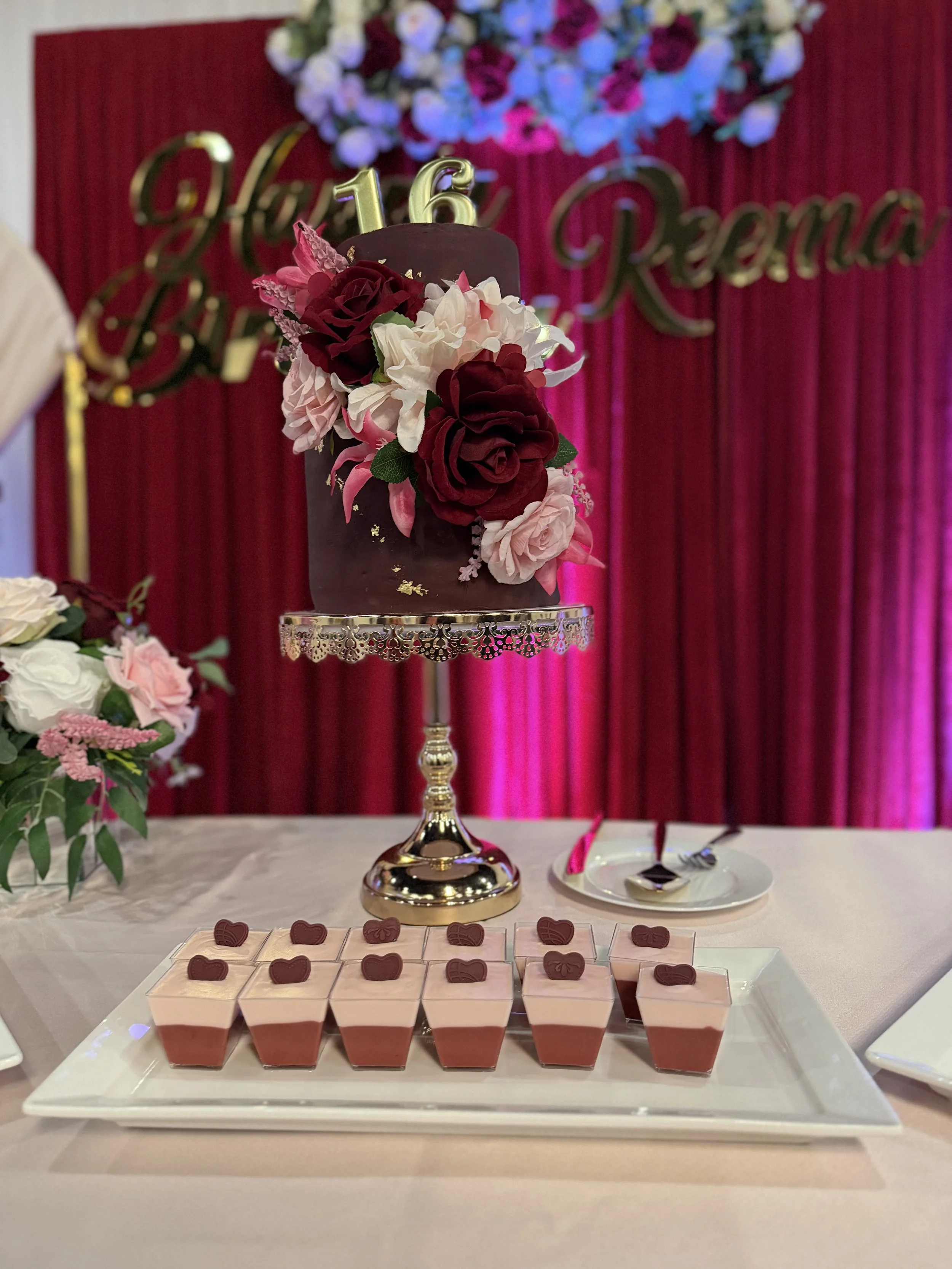 A decorated cake with flowers and a number 16 topper, set on a stand in front of a red curtain background, with a tray of small desserts topped with chocolate hearts.
