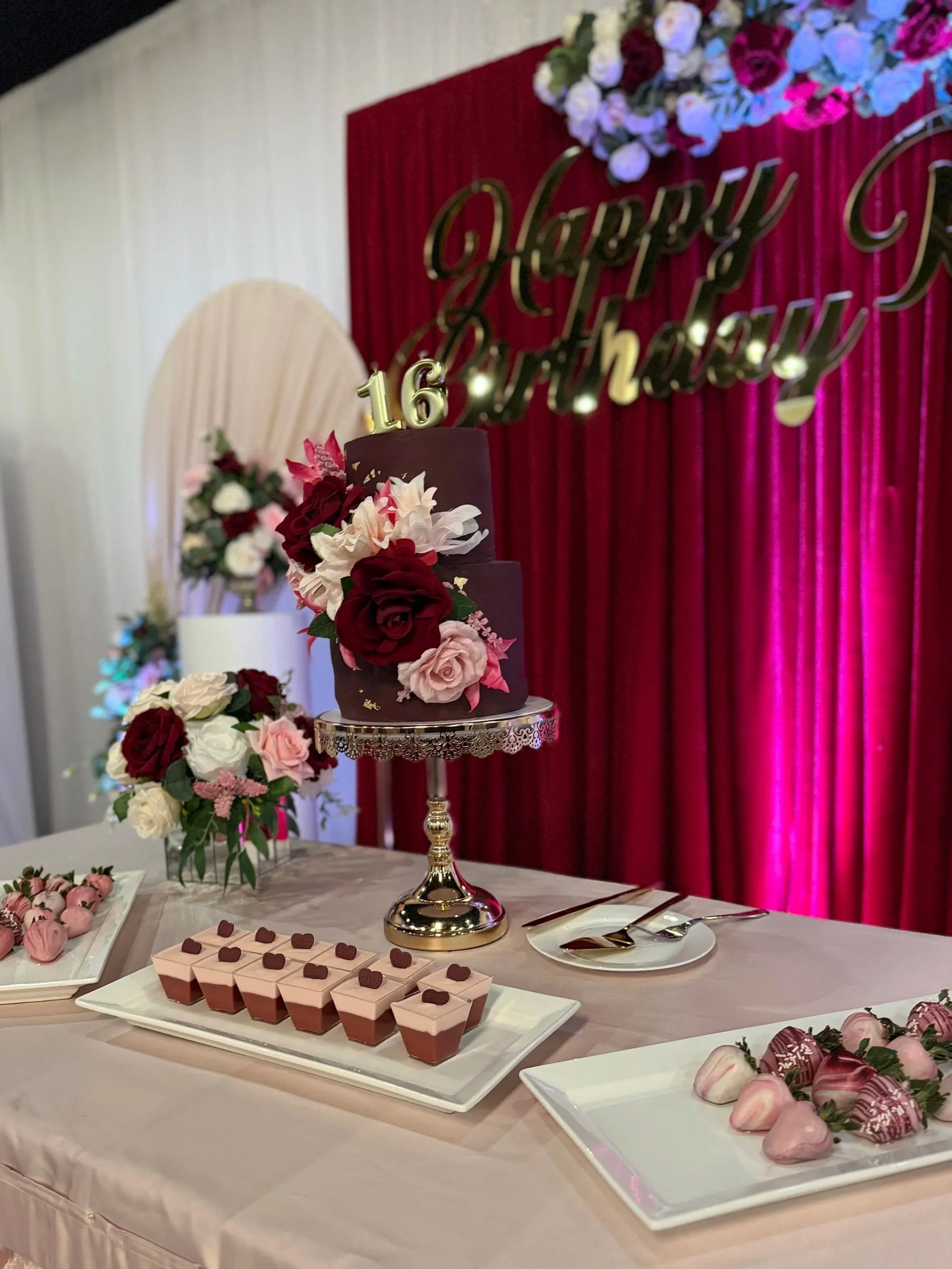 A dessert table decorated with flowers and chocolates, featuring a two-tier chocolate cake with flowers and a 'Happy Birthday' banner in the background.