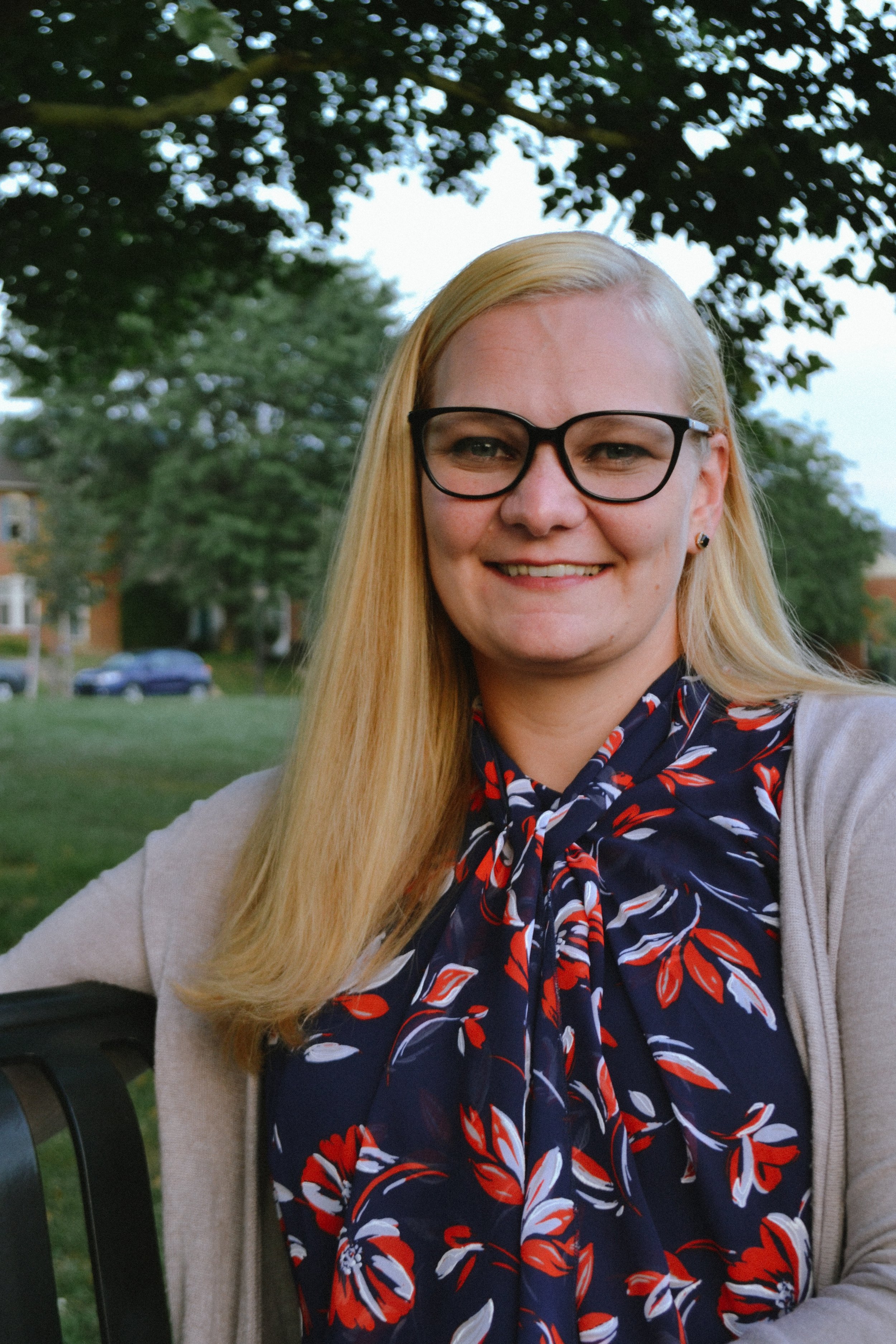 Person with blonde hair, glasses, and a floral-patterned blouse, sitting outside on a bench with trees in the background.