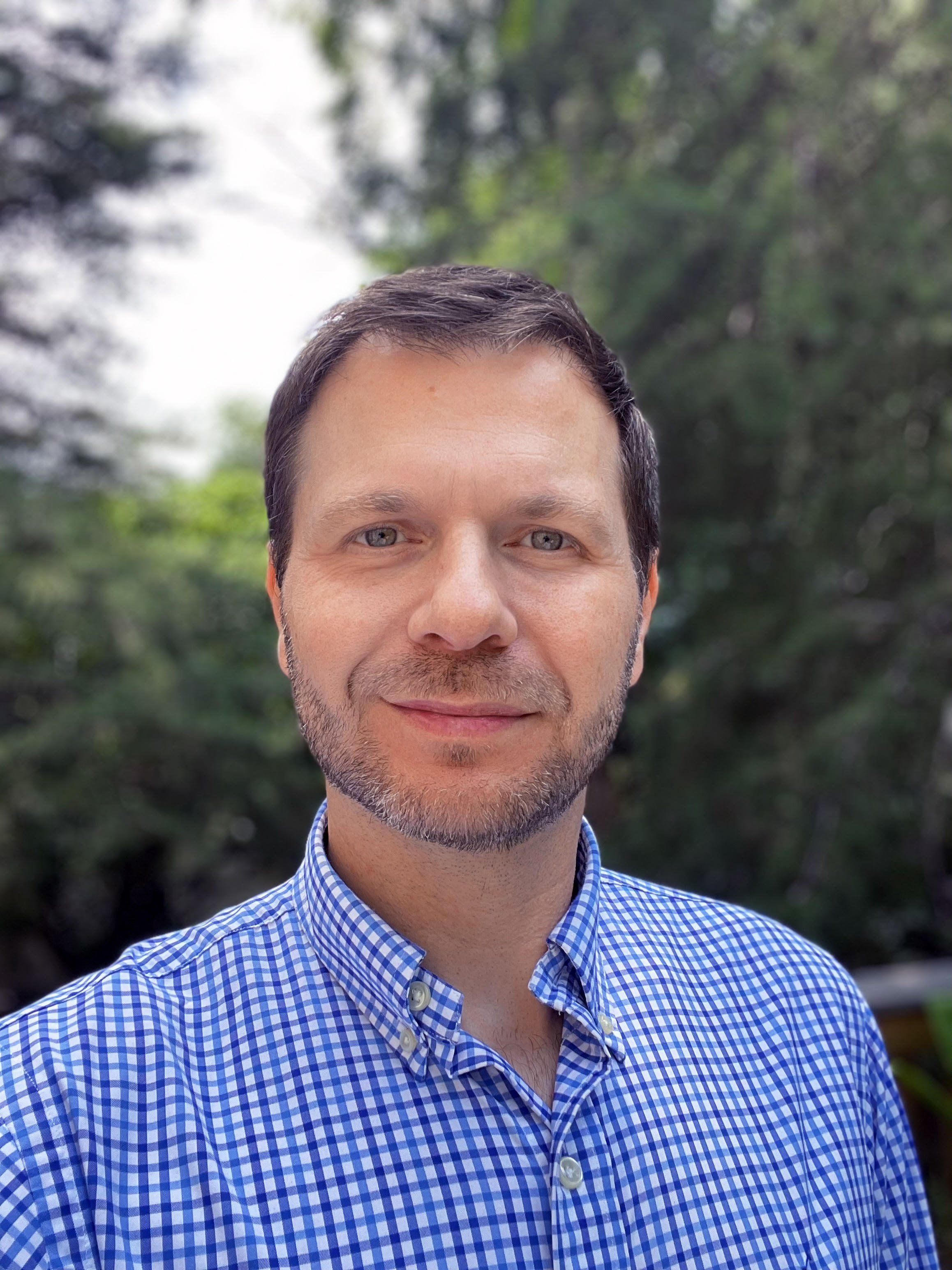 Man in blue checkered shirt outdoors with trees in background.