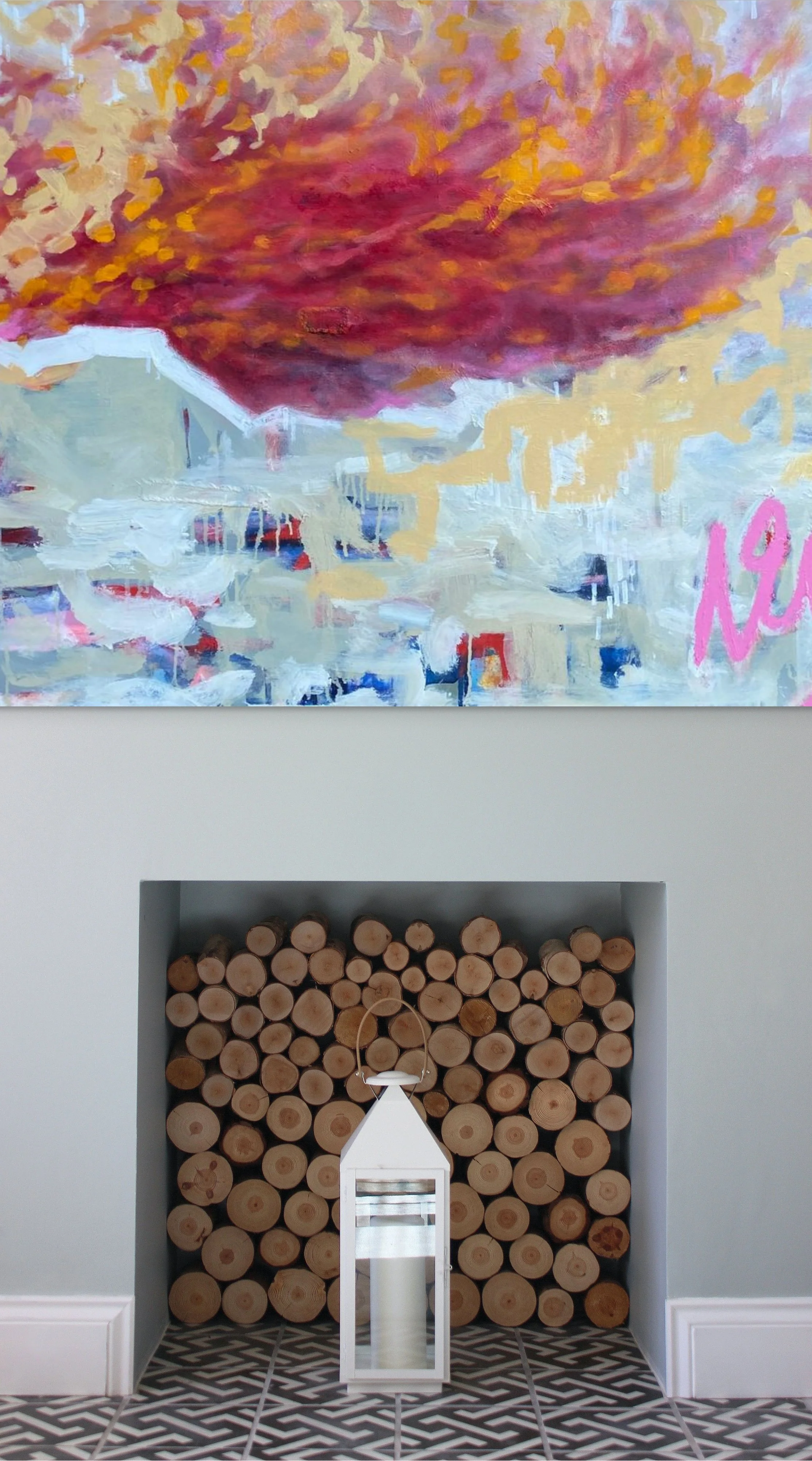 An abstract painting with a large, reddish-orange cloud-like shape at the top and a colorful, blurred cityscape below. The painting has the word 'Note' in pink at the bottom right. Below the painting, there is a neatly stacked pile of cut logs within a recess in the wall, and a white lantern with a candle inside is placed on the floor in front of the logs.