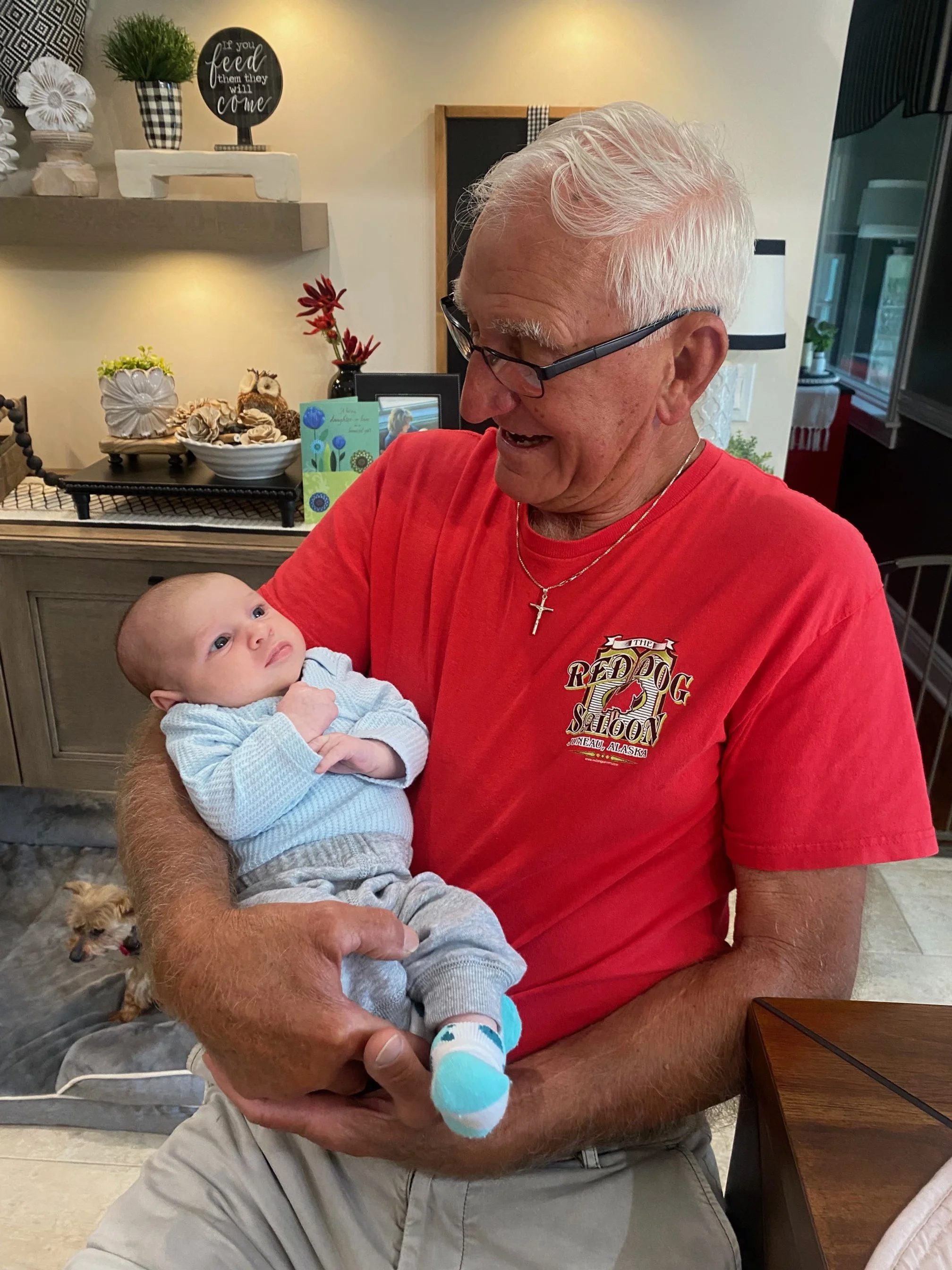 An elderly man holding a baby in a home setting. The man is smiling and wearing glasses and a red T-shirt with a logo. The baby is looking up at the man, wearing a light blue sweater, grey pants, and colorful socks. In the background, there are decorative items on shelves, a lamp, and a small dog lying on a blanket.
