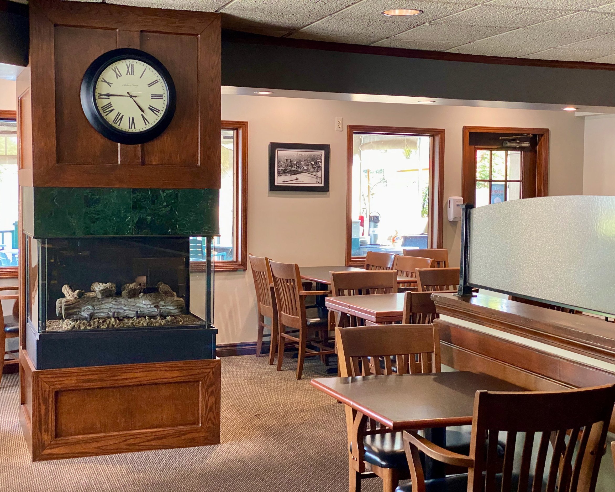 Interior of a restaurant or cafe with wooden tables and chairs, a fireplace with a clock on a wooden mantel, and windows letting in natural light.