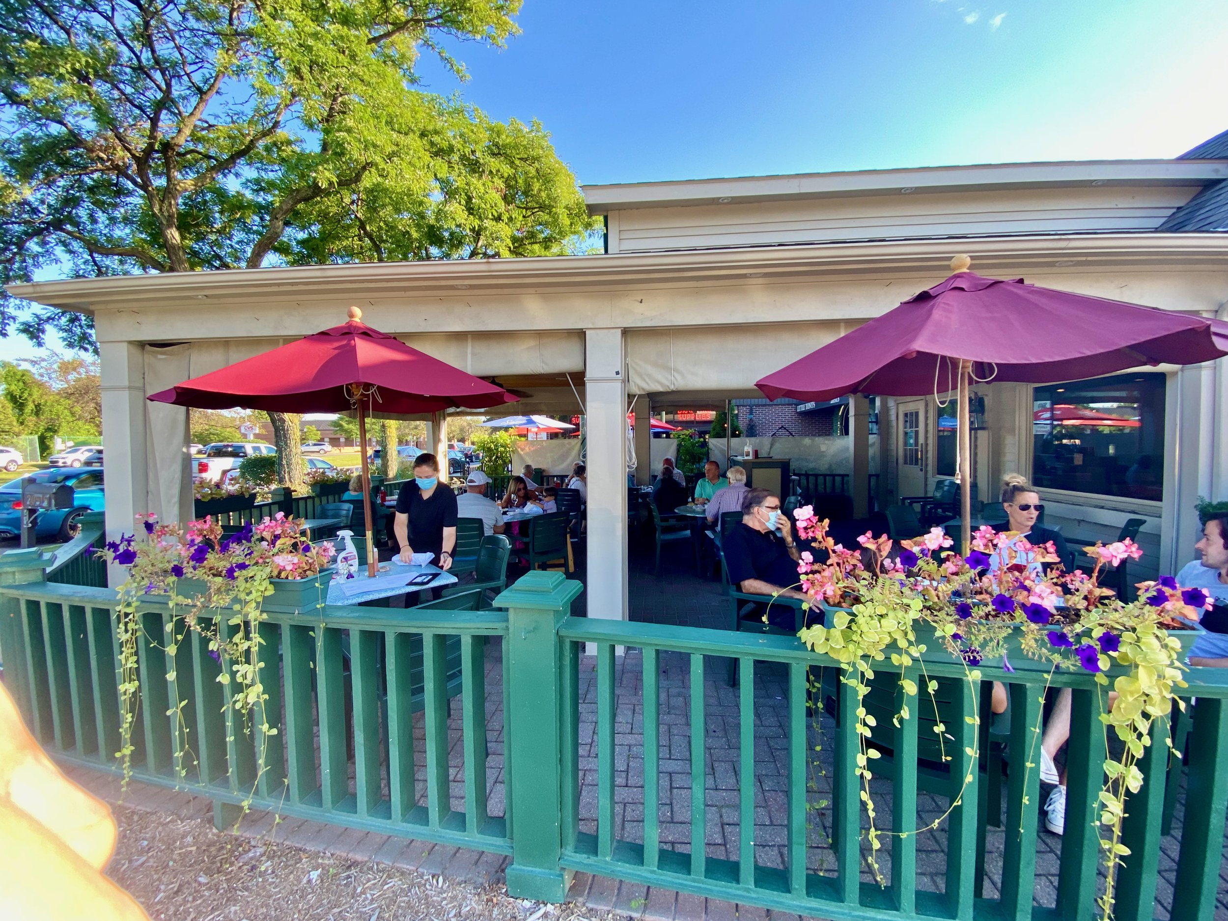 Outdoor patio scene at a restaurant or cafe with several people seated at tables under large purple umbrellas. There is a green railing with purple and pink flowers in the foreground. The background features a building with white siding, a large tree with green leaves, and a parking lot with cars. The sky is clear and blue.
