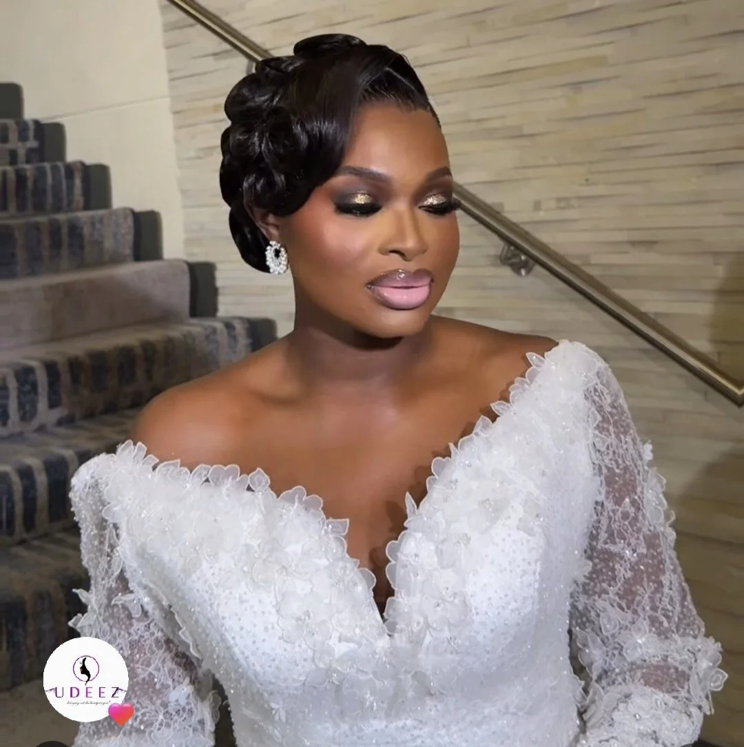 A woman in an elegant white lace off-shoulder dress with detailed embroidery, gray makeup eyeshadow, pink lipstick, and large dangling earrings, standing on a staircase with a wood-paneled wall background. Makeup and hair styled by House of Udeez.