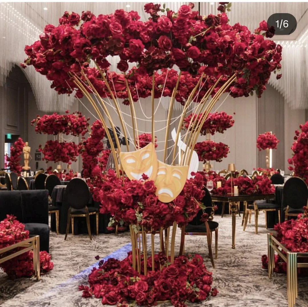 Elegant event space decorated with large arrangements of red roses and gold theatrical masks, featuring bold floral centerpieces and black chairs, designed for a celebration by stylist vendor TP Collective.