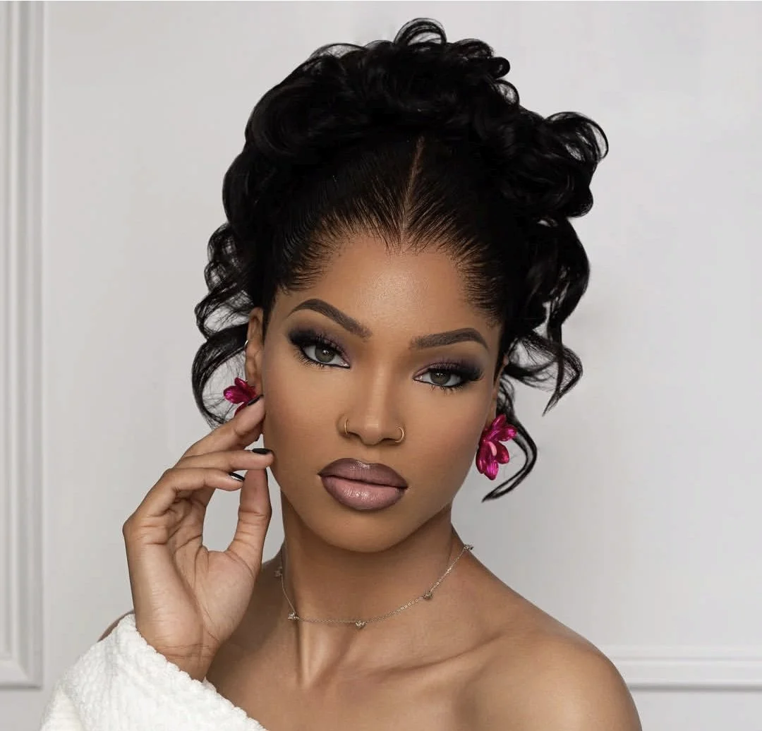 Portrait of a woman with styled curly hair, wearing makeup, pink earrings, and a necklace, against a plain background. Styling by makeup and hair artist: Leemah.
