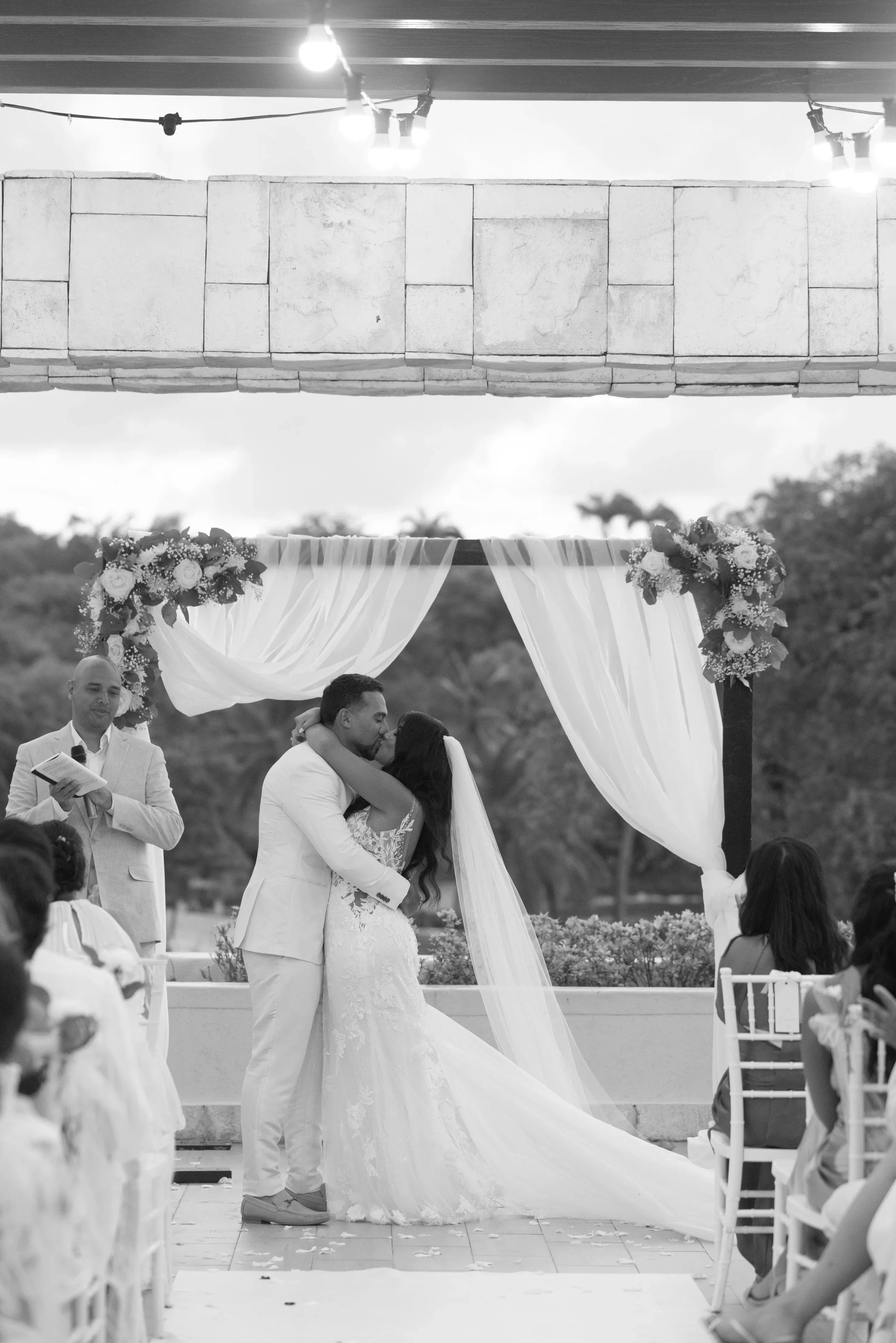 A black and white photo of a wedding ceremony with a bride and groom kissing under an arch decorated with flowers and draped fabric, with wedding guests seated on either side.