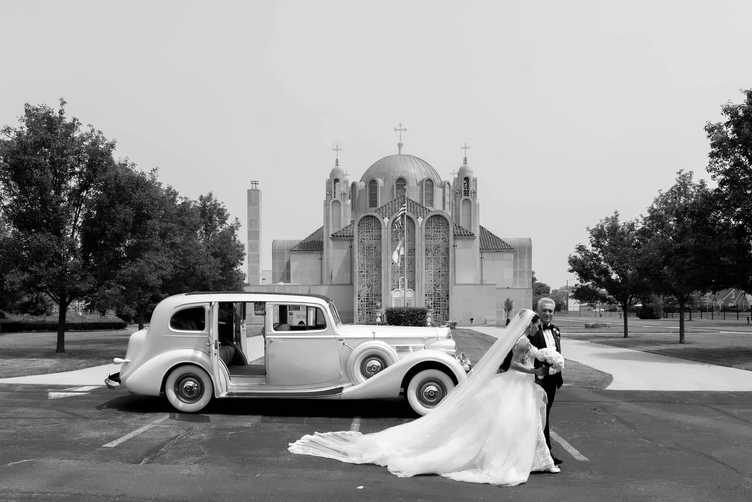 A vintage white car parked in front of a church with a large dome and three crosses on top. A bride and groom are standing beside the car, with the bride holding a bouquet and the groom in a tuxedo. The bride's dress train is spread on the ground. Trees and people are in the background.