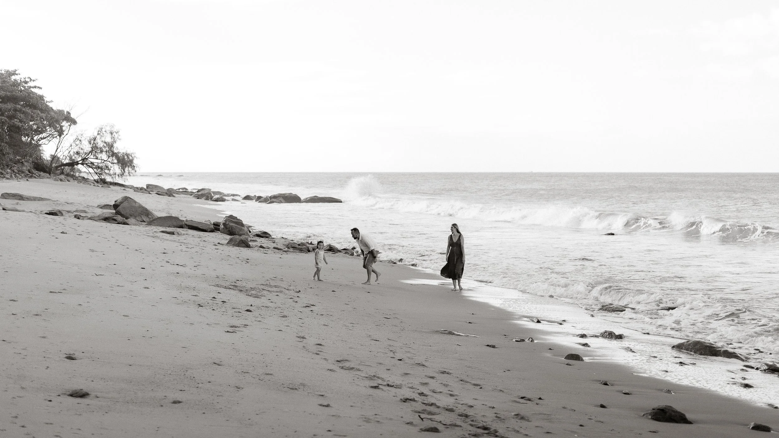 A black and white photo of three people, two adults and one child, walking on Ellis Beach near the water's edge, with rocks and trees on the left side and ocean waves in the background.