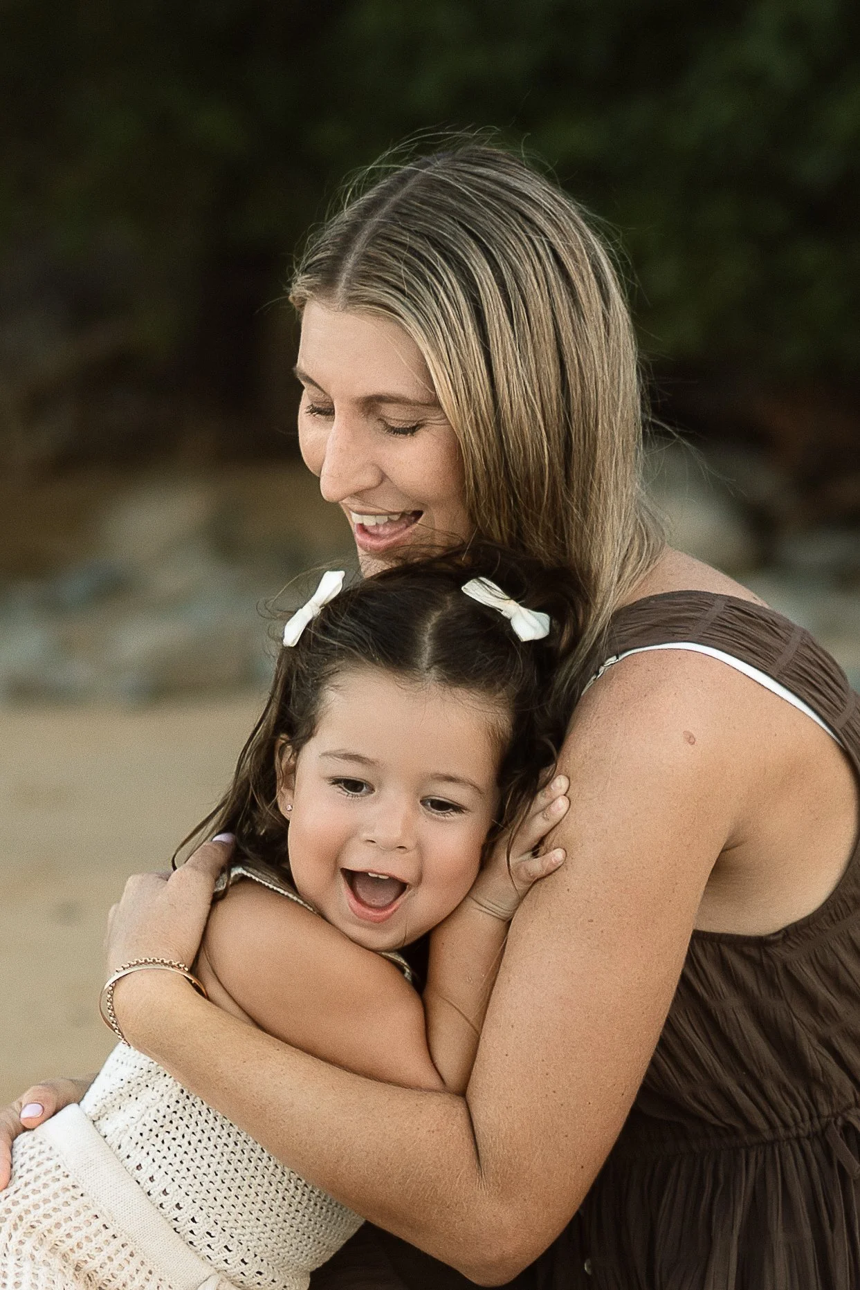 A woman with blonde hair hugging a young girl with brown hair, both smiling and showing joy in an outdoor setting.