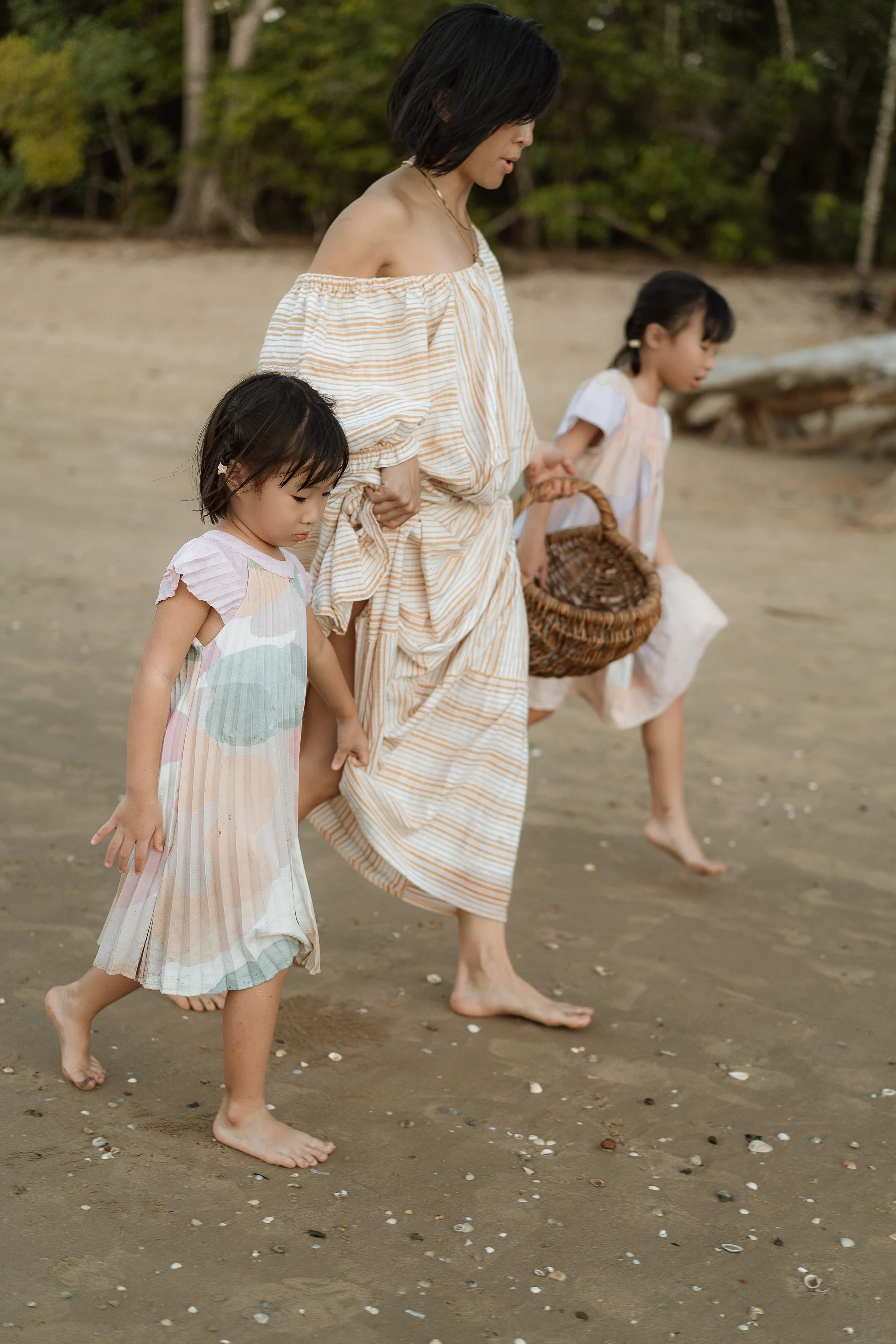 A woman with short black hair in an off-the-shoulder beige-striped dress walking barefoot on a sandy beach accompanied by two young girls also barefoot, one wearing a pastel striped dress and the other in a light-colored dress, with trees and greener