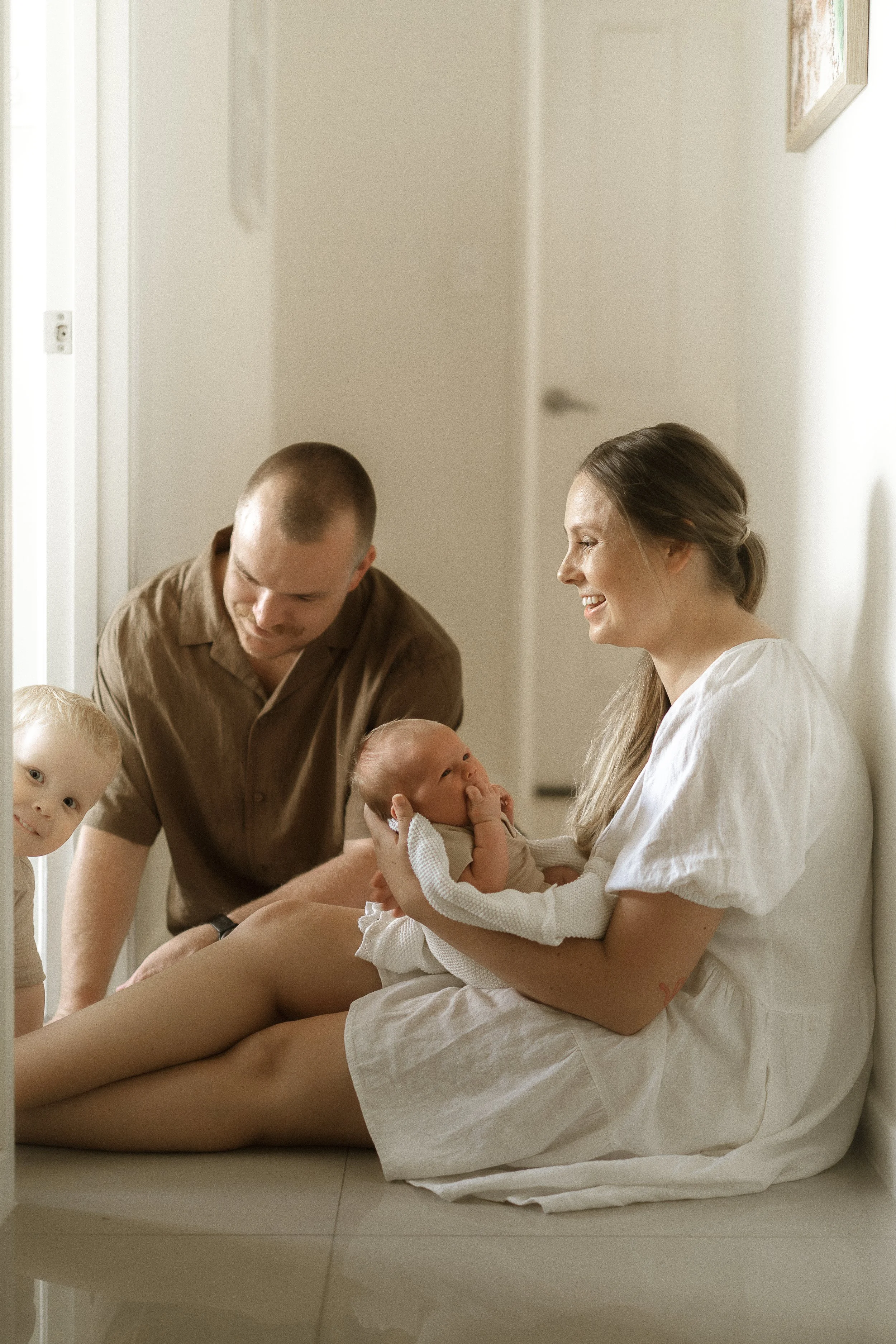 A woman sitting on the floor holding a baby, smiling at a man who is leaning in towards them. Two young children are nearby in a bright, neutral-colored room. Cairns newborn home session.