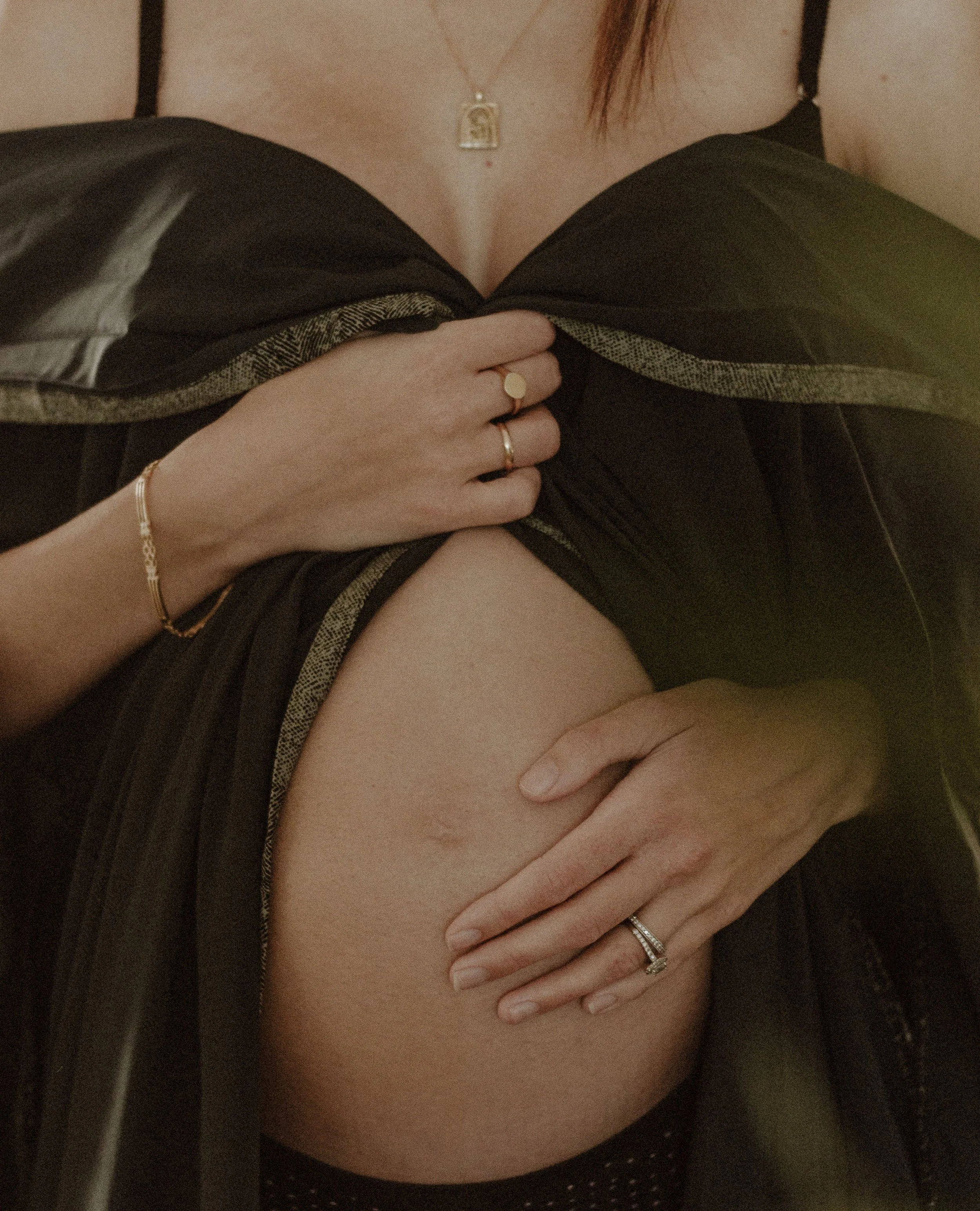 A person holding their breast with one hand, wearing rings, bracelets, and a necklace with a religious pendant. The person is dressed in black lingerie and a satin robe, with the bra strap visible. Palm Cove maternity home session.