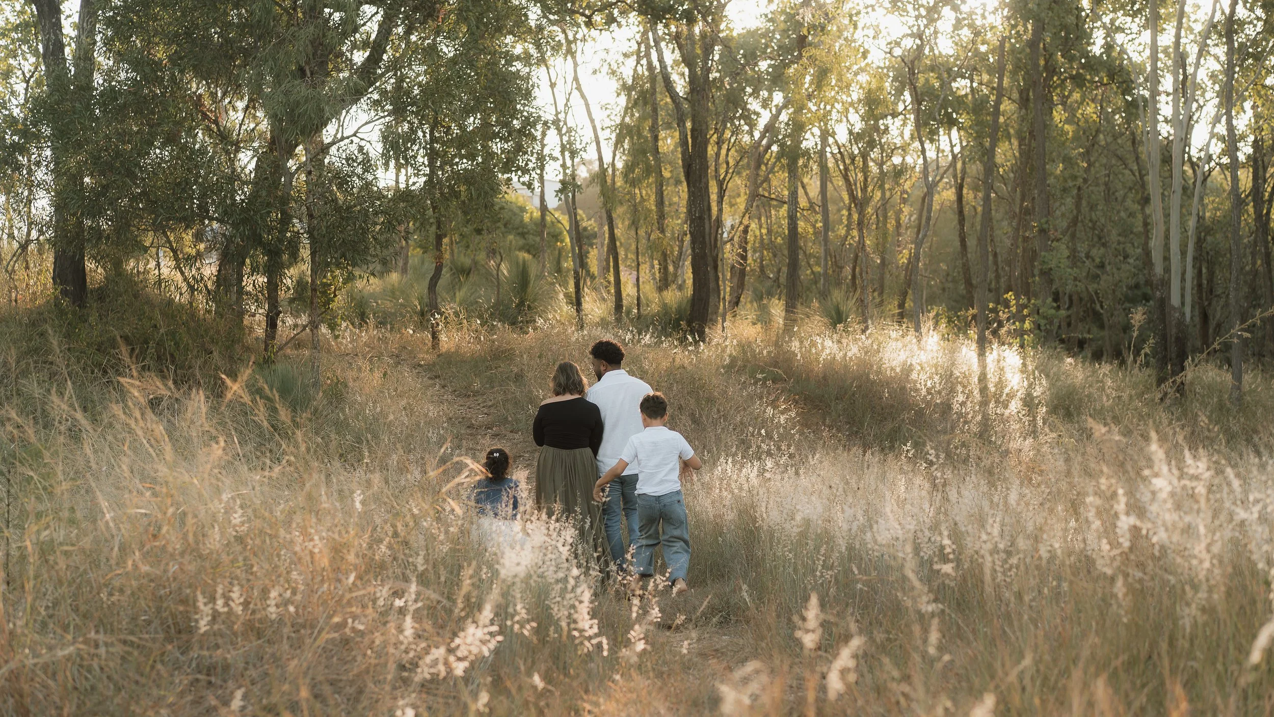 A family walking along a dirt path through a lush, green forest in the late afternoon or early evening.