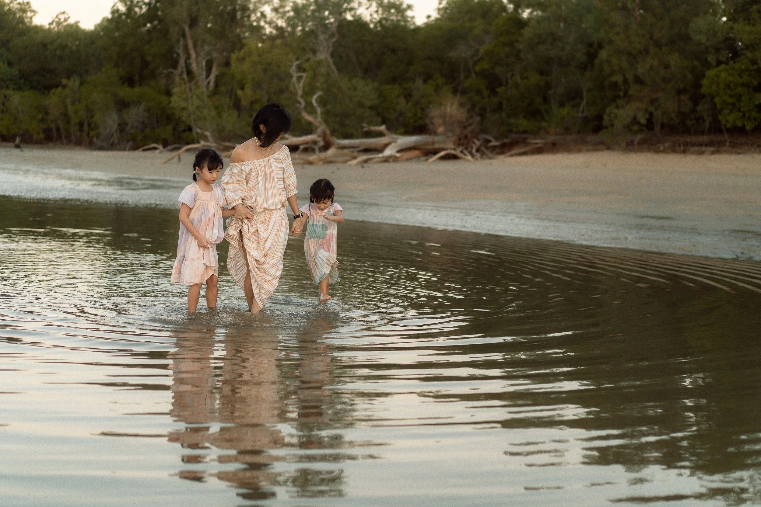 A woman walking with two young girls along the shoreline of a beach during sunset or early evening, with trees in the background, water reflections, and driftwood on Kewarra beach.