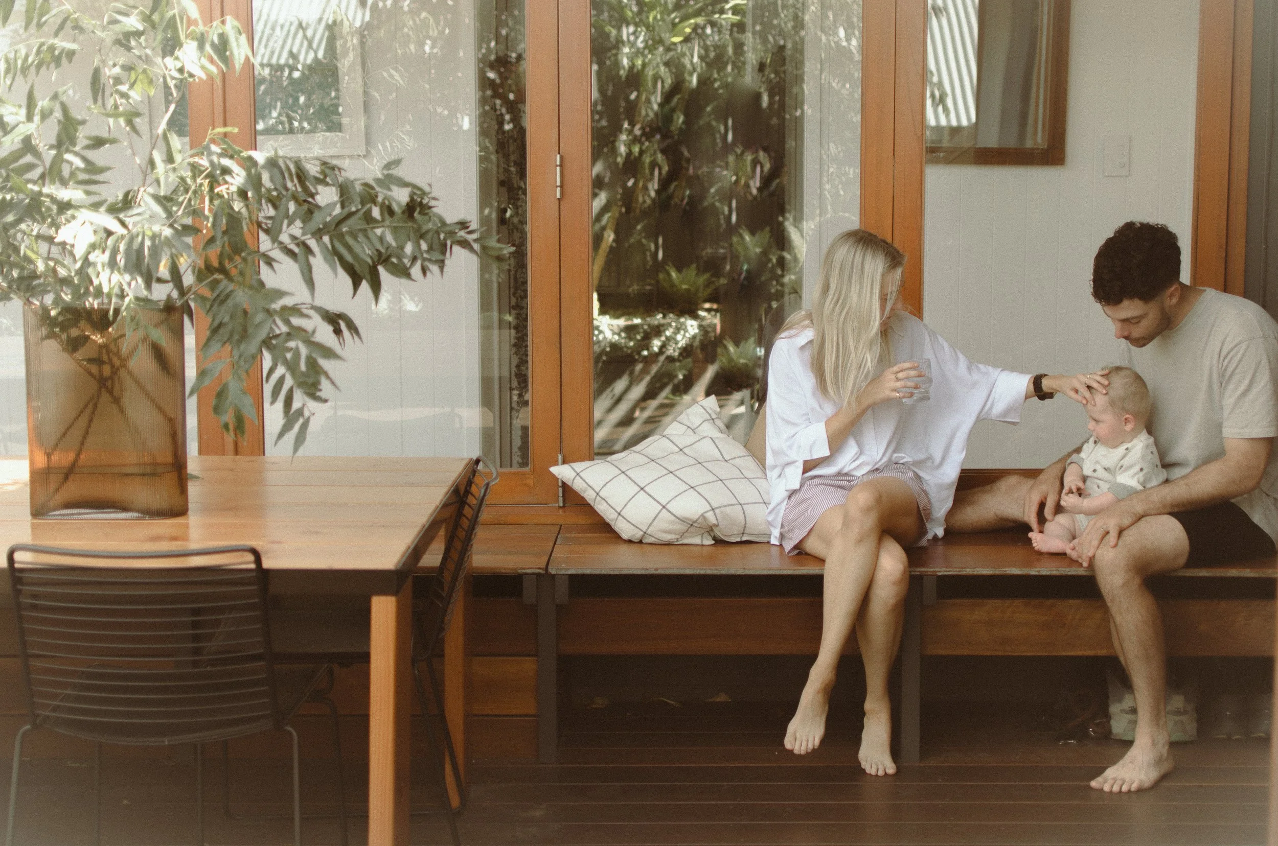 Family sitting on wooden deck near large glass doors with plants outside, woman and man with a toddler in an indoor-outdoor space. Palm Cove home session.