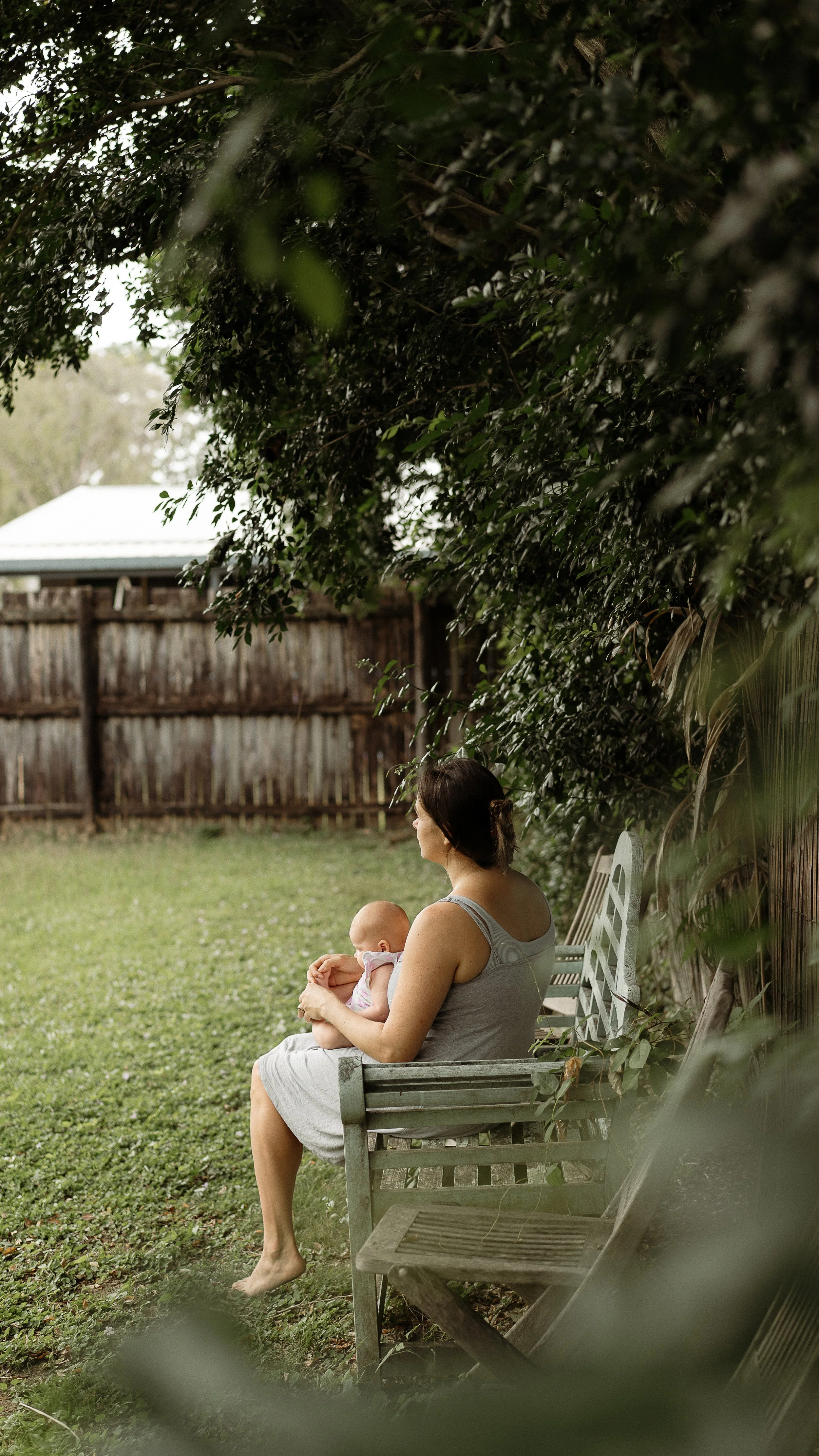 A woman sitting on a park bench holding a baby in her lap, with a backyard and fence in the background, surrounded by greenery.