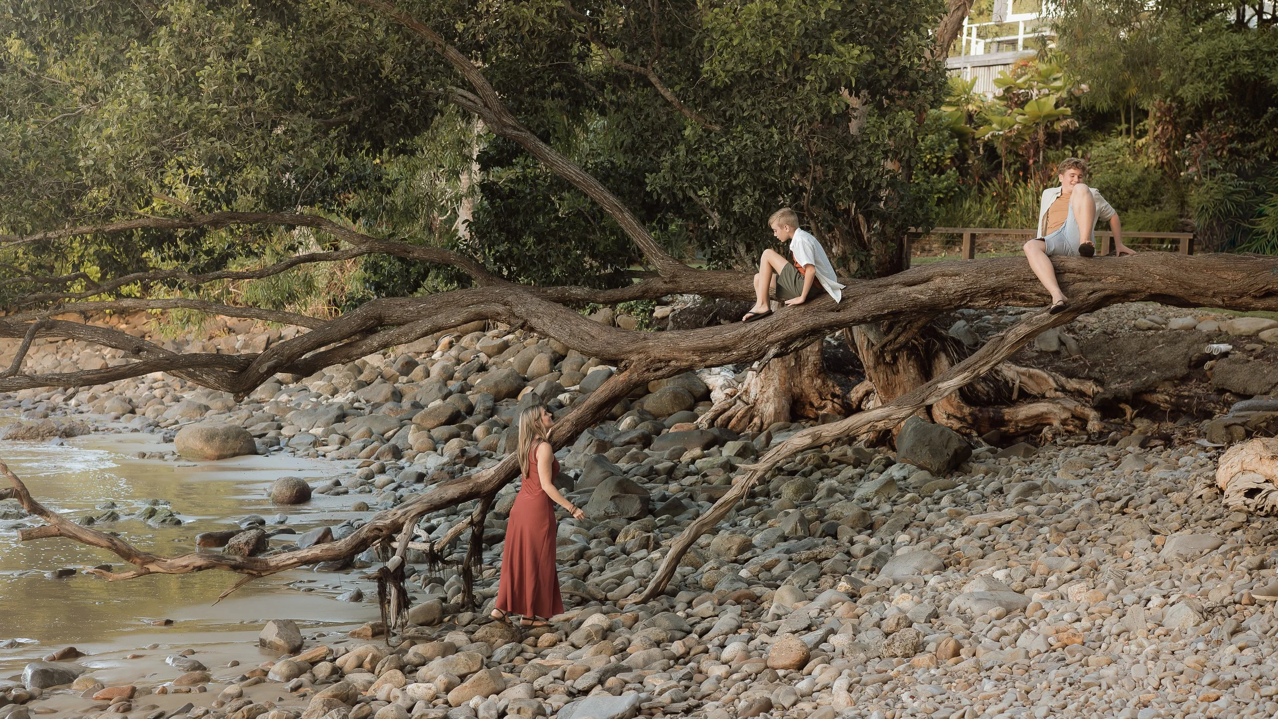 Three children playing on a fallen tree across a rocky riverbank with greenery in the background. Port Douglas family session.