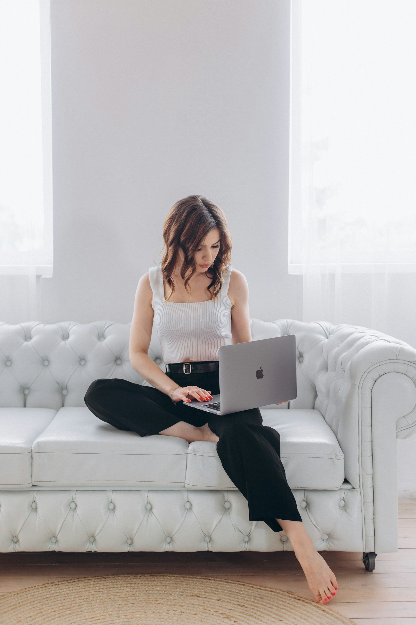 Woman sitting cross-legged on a white tufted sofa working on a silver MacBook, looking up virtual therapy  in a bright room with white walls and large windows.