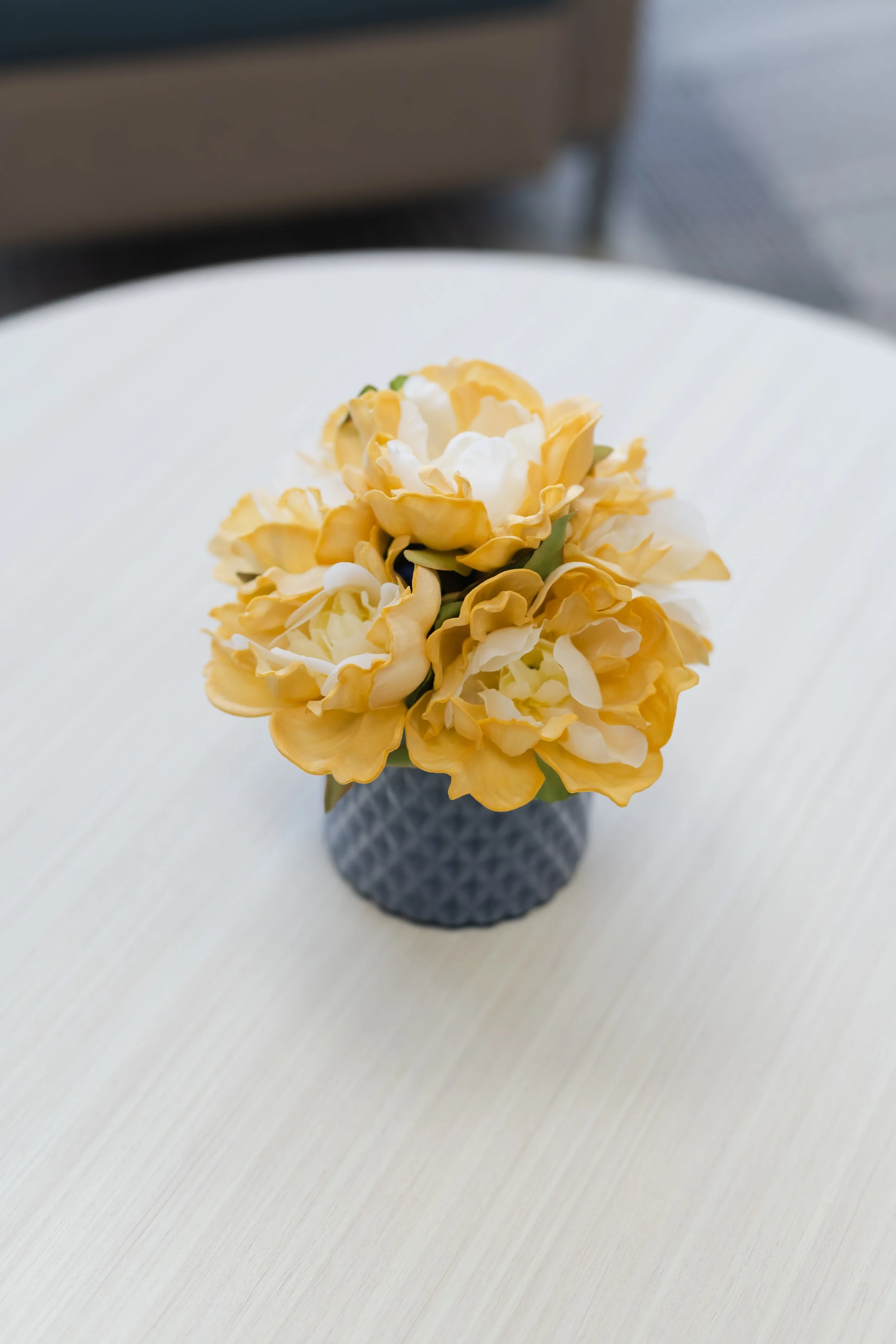 A small bouquet of yellow and white artificial flowers in a dark patterned vase on a white table.