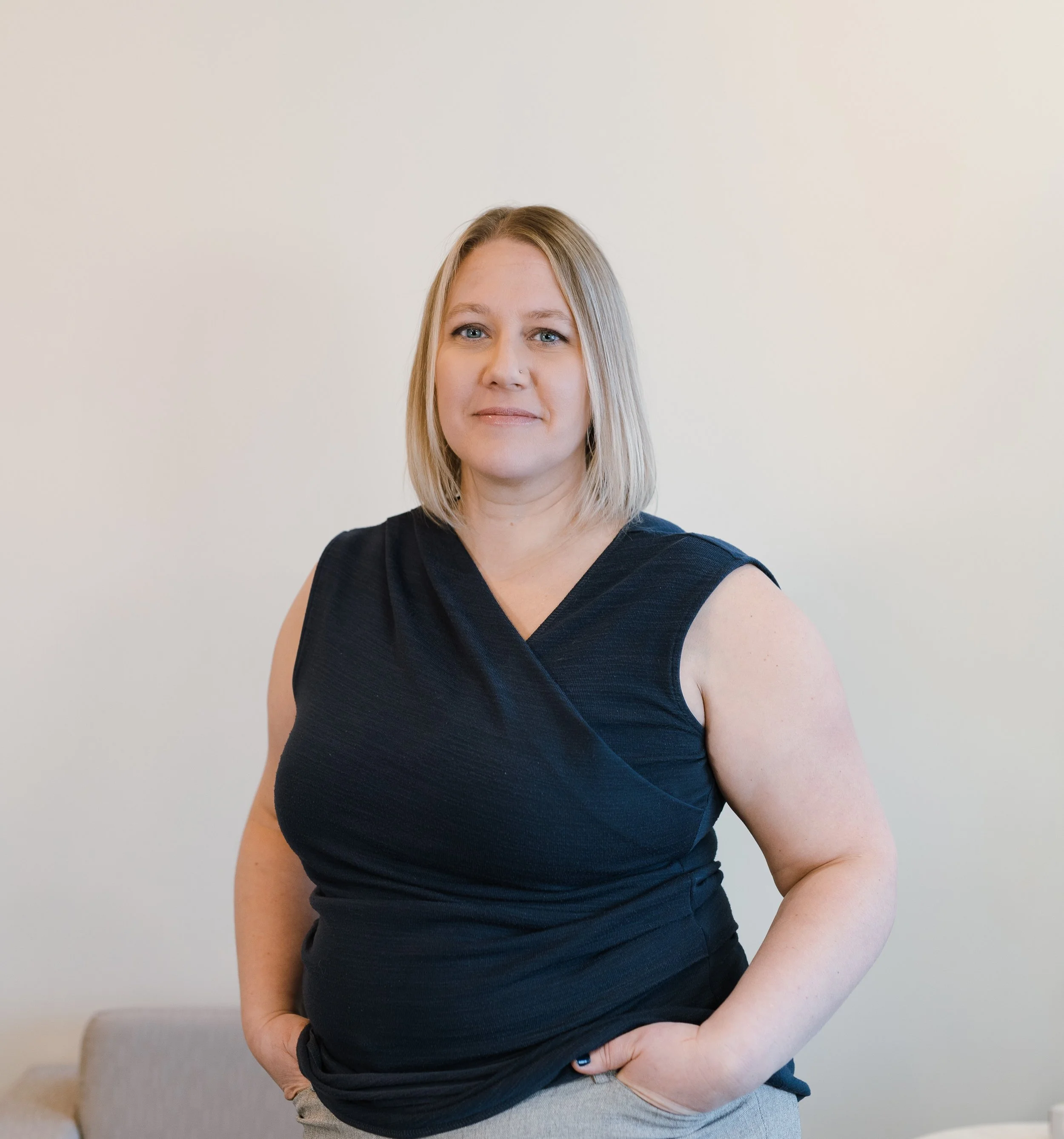 A woman with shoulder-length blonde hair wearing a black sleeveless top and gray pants, standing against a plain light-colored wall.