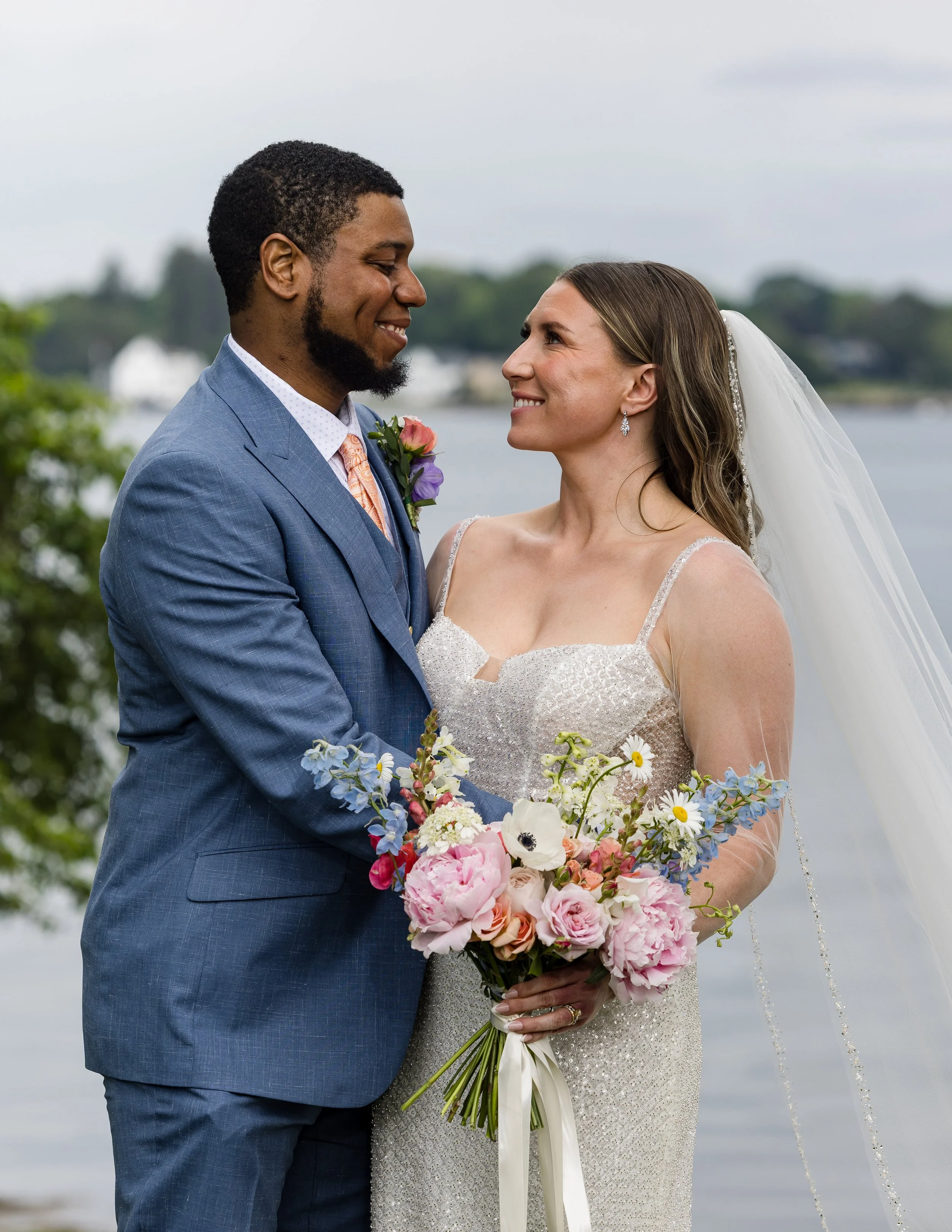A newlywed couple standing outdoors by a body of water, gazing into each other's eyes, with greenery in the background. The bride is holding a bouquet of pink, white, and purple flowers, wearing a wedding dress and veil. The groom is dressed in a blue suit with a boutonnière.