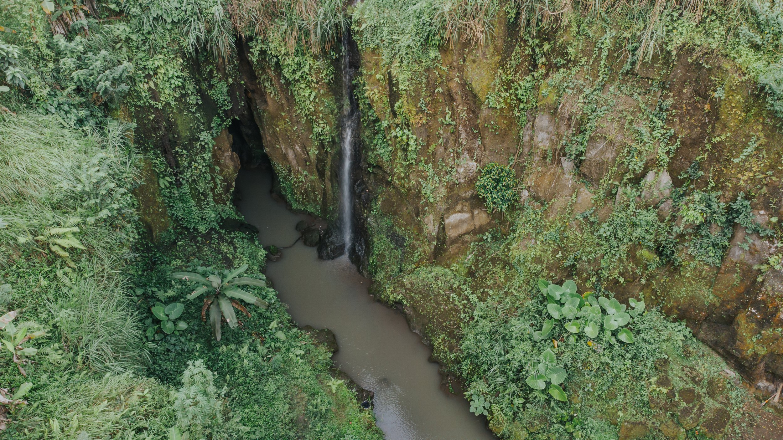 A small waterfall flowing into a river surrounded by dense green tropical vegetation. High-end retreat center in Pejeng, Ubud, Bali