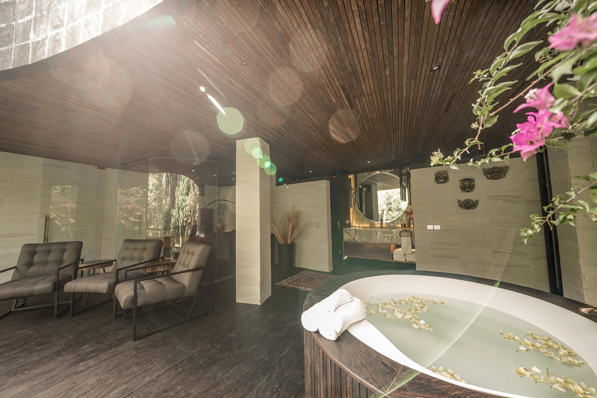 Modern interior with dark wooden ceiling, white walls, and modern furniture including lounge chairs. There are decorative masks on the wall and a partially visible bathtub with flowers and towels nearby, indicating a spa or luxurious home setting.