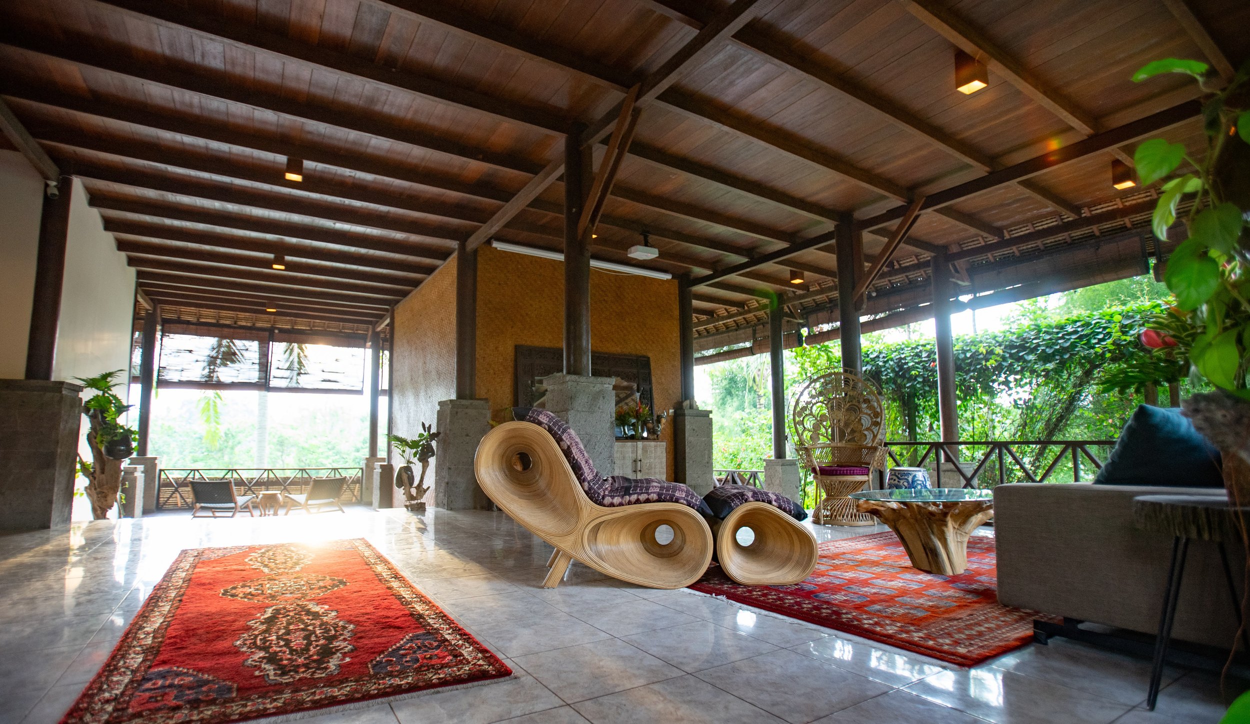 Indoor living space with wooden beams on ceiling, large windows, and tropical plants. Features a red patterned rug, lounge chairs, rattan chairs, and various decorative elements.