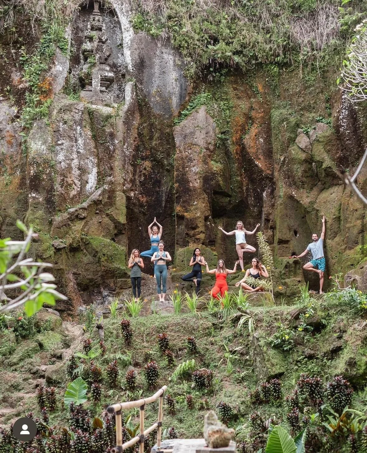 People practicing yoga outdoors in front of a rocky, moss-covered cliff with lush greenery in Bali, Pejeng, Kelebutan