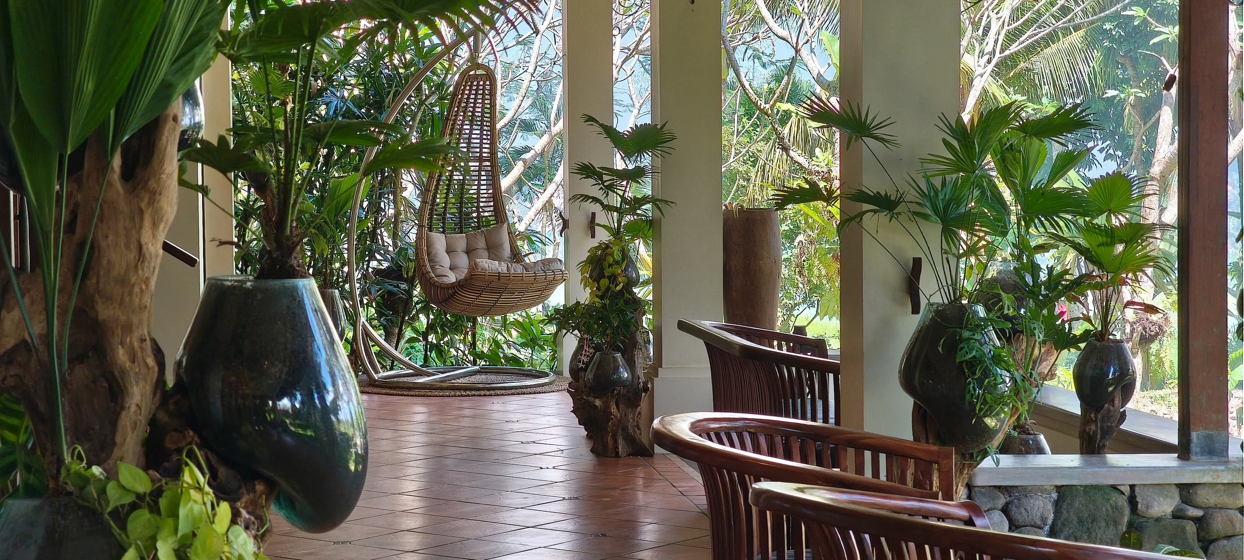 Lush indoor space with large green plants in vases, wooden chairs, and a hanging wicker chair by large windows with trees outside. High-end retreat center in Pejeng, Ubud, Bali