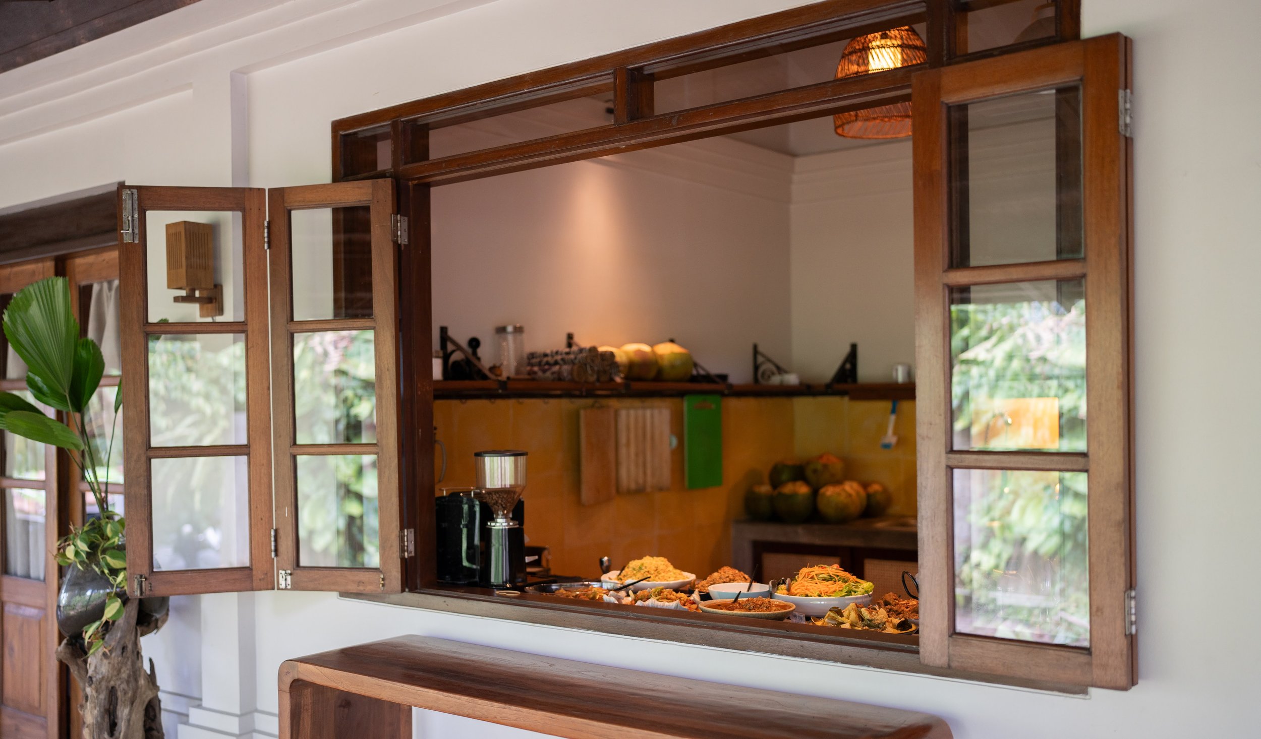 A view through a wooden window in a wall shows a kitchen with a counter, some produce, and a hanging woven lamp.High-end retreat center in Pejeng, Ubud, Bali