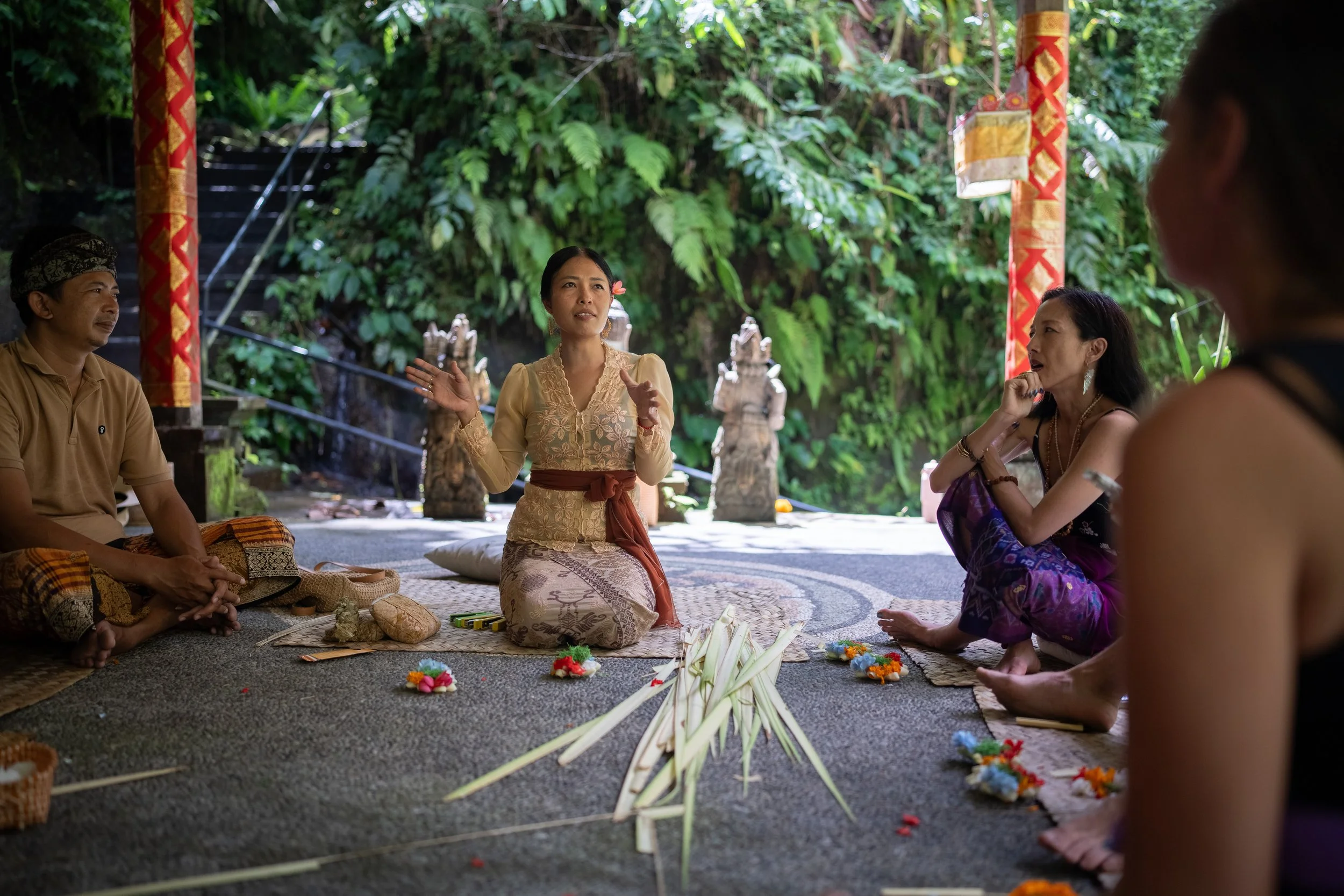 A group of people, including a woman in traditional attire, gathered outdoors in a lush, green natural setting with statues in the background, participating in a cultural ceremony or discussion. High-end retreat center in Pejeng, Ubud, Bali
