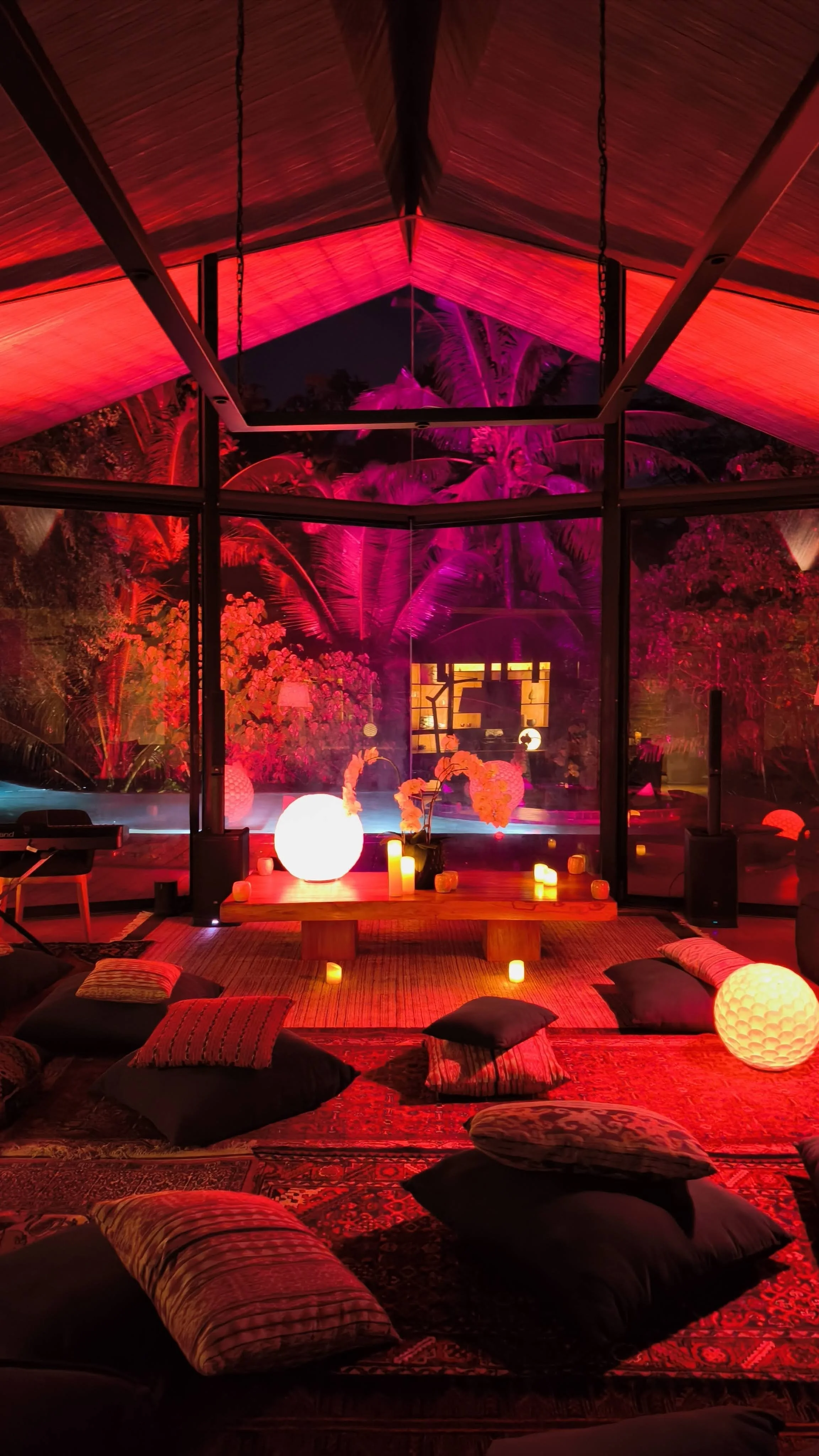 A cozy indoor space with cushions and pillows on the floor, illuminated by various candles and decorative lamps. Large glass windows reveal a lush outdoor garden at night, illuminated with pink and purple lighting, and tropical plants are visible outside.