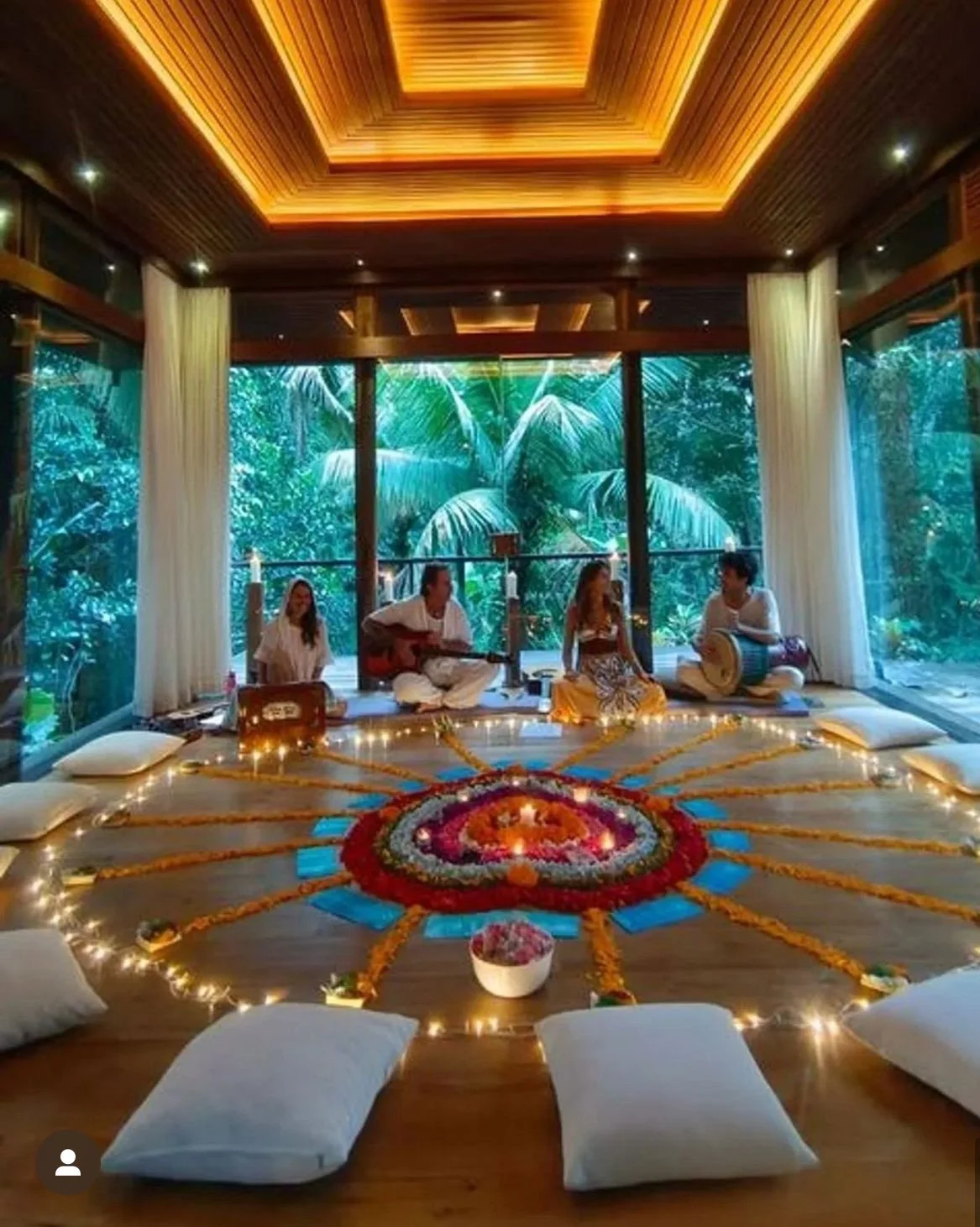 A group of four people sitting on a carpeted floor inside a room with large glass windows overlooking lush green tropical trees. They are surrounded by candles and flowers arranged in a circular pattern with blue cards, pillows, and candles, possibly