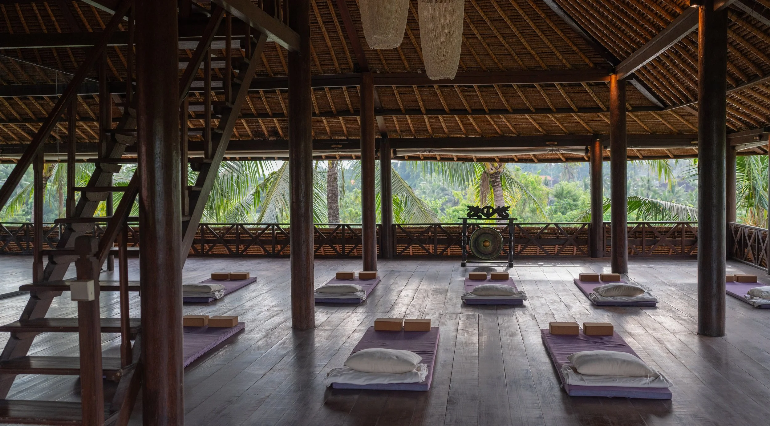 Indoor yoga space with mats and cushions, wooden structure, surrounded by greenery visible through open sides. High-end retreat center in Pejeng, Ubud, Bali