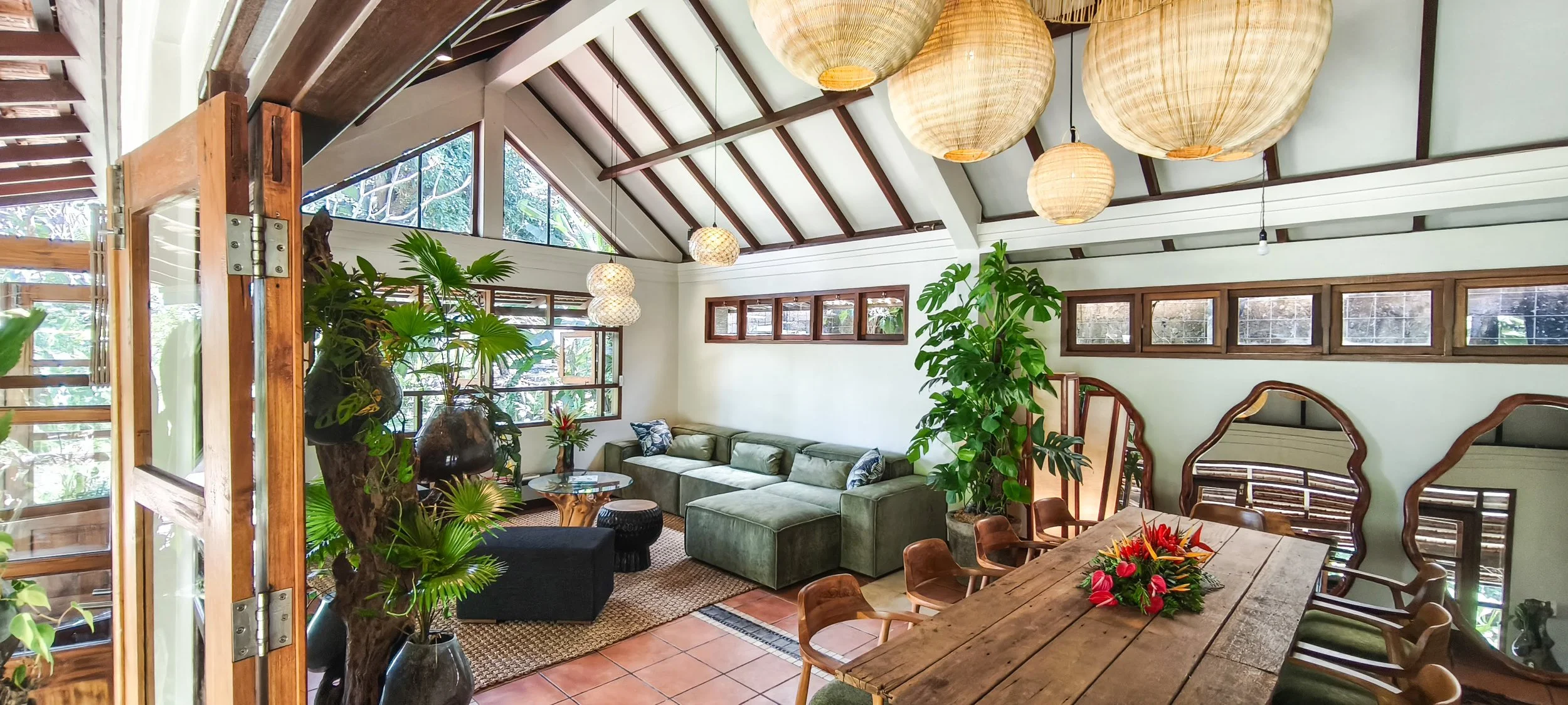 A spacious indoor living room with large windows, green plants, a green sectional sofa, wooden furniture, hanging paper lanterns, and a wooden dining table with a floral centerpiece. High-end retreat center in Pejeng, Ubud, Bali