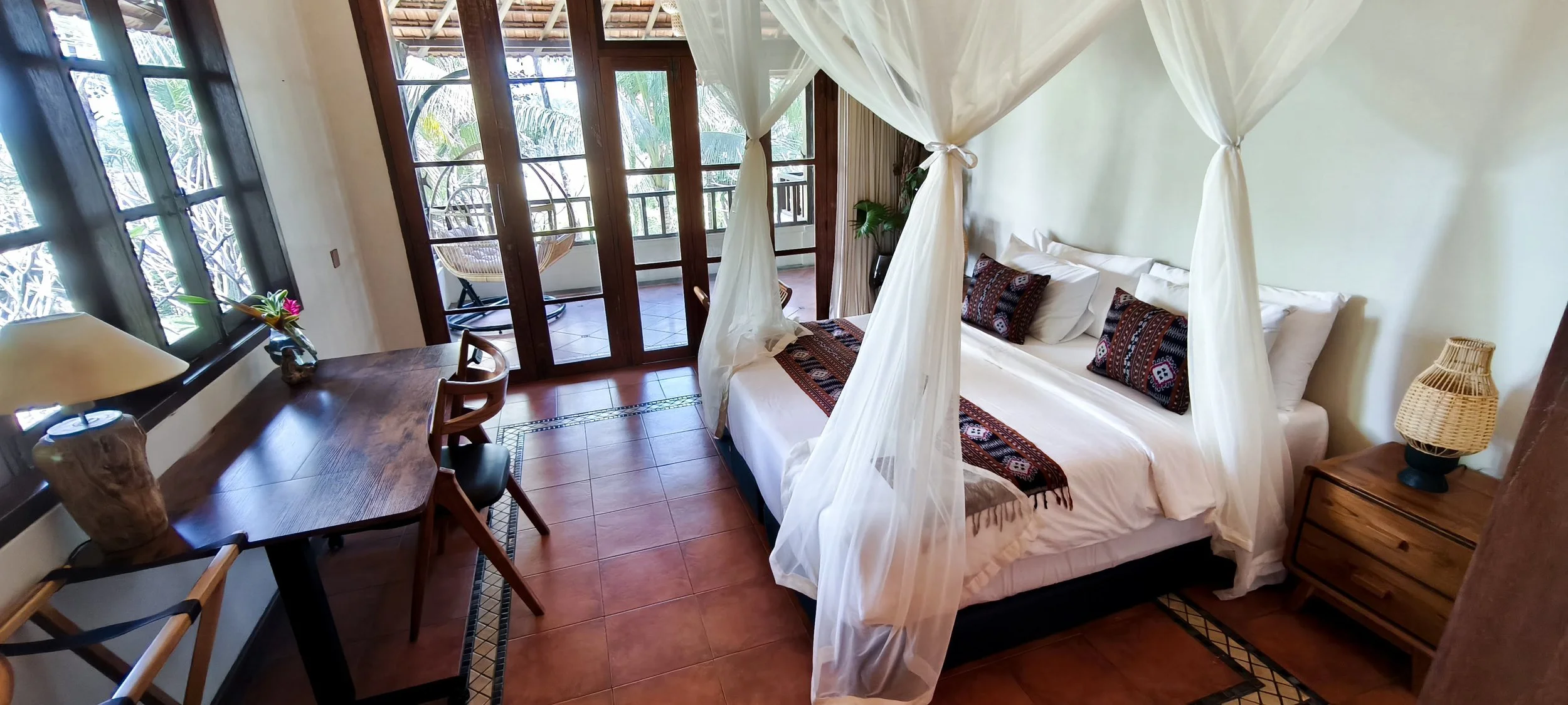 A cozy bedroom with a four-poster bed draped with white curtains, decorated with patterned pillows and a woven runner. The room has a wooden nightstand with a lamp, a small desk with chairs, and a balcony visible through glass doors, with outdoor seating and greenery.
