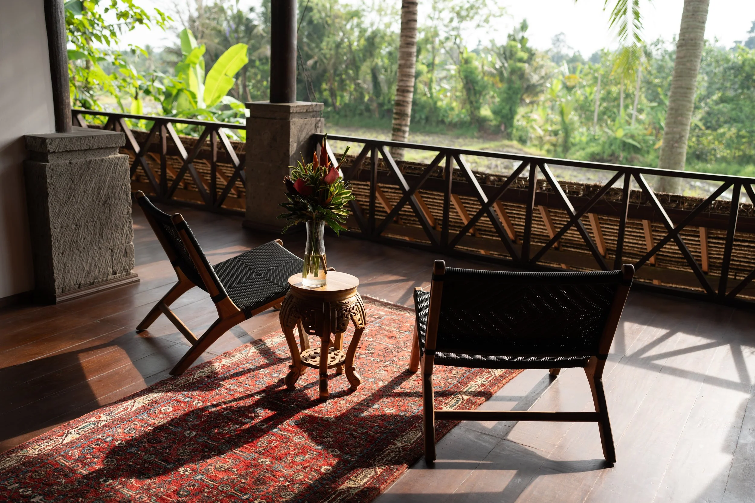 A porch with wooden railings overlooking lush green trees and palm trees in the background. There are two black chairs, a small carved wooden table with a floral arrangement in a glass vase, and a red patterned rug on a wooden floor.