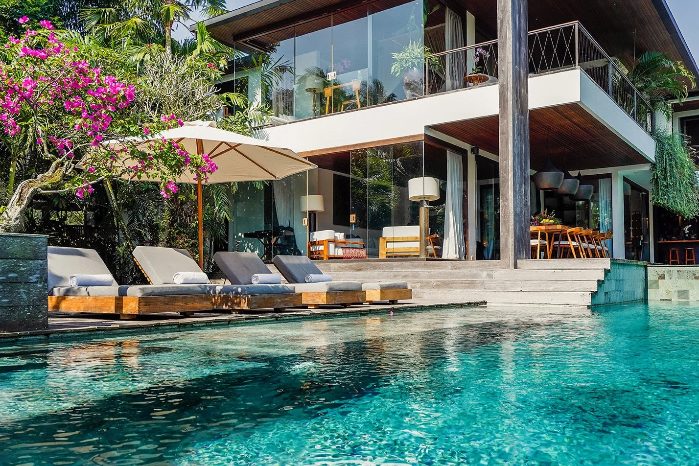 Modern house with glass walls overlooking a swimming pool with lounge chairs and an umbrella. High-end retreat center in Pejeng, Ubud, Bali