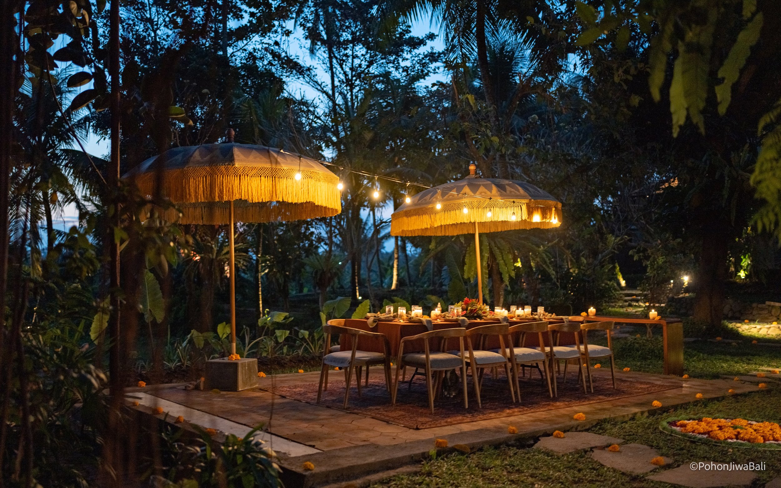 An outdoor dining setup at dusk with a long table surrounded by chairs, lit by candles and decorated with flowers. Overhead umbrellas with hanging lights and lush greenery and trees in the background. High-end retreat center in Pejeng, Ubud, Bali