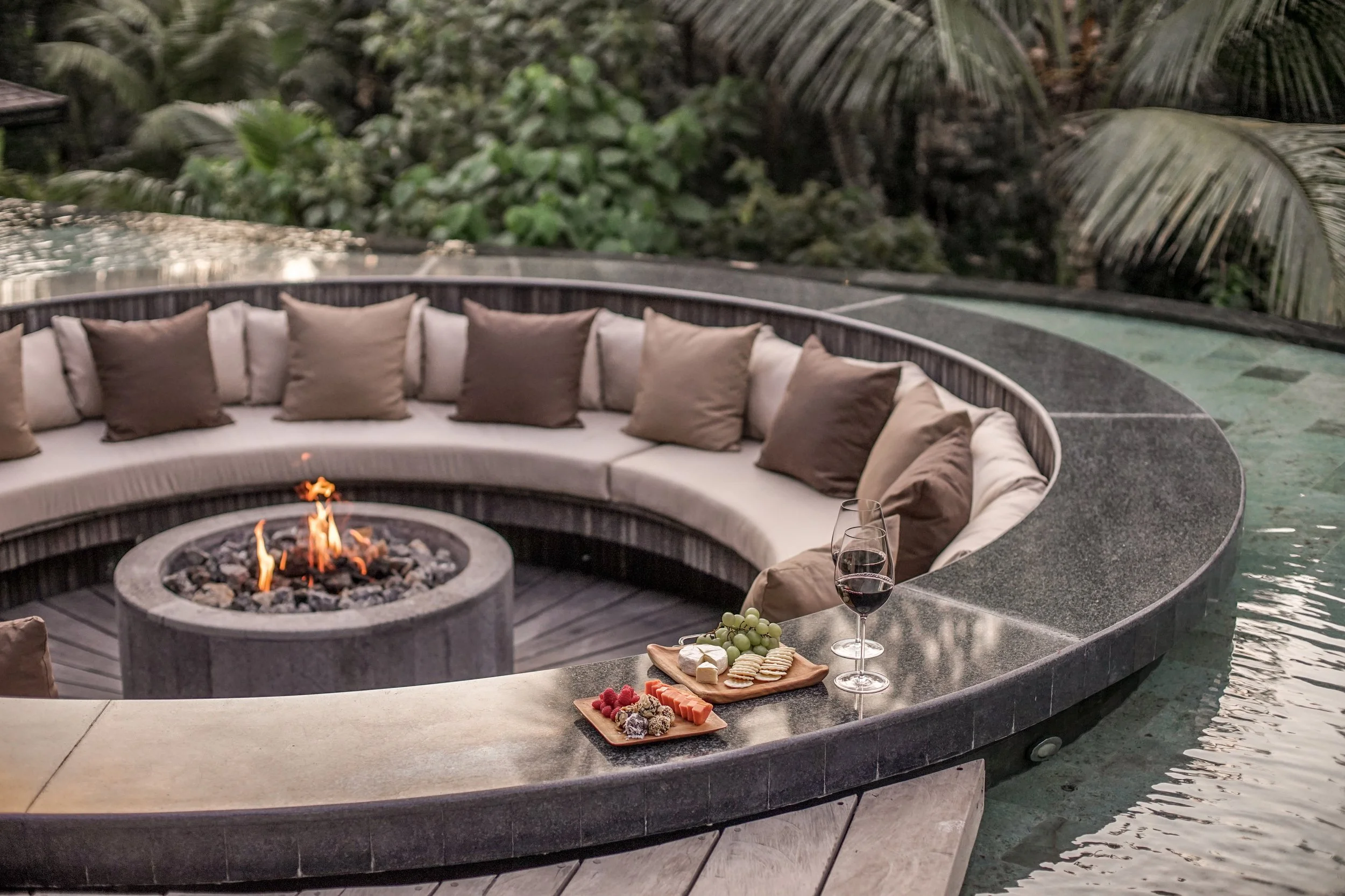 Outdoor seating area with a circular fire pit and beige cushions around it, surrounded by lush greenery and large palm leaves, with a tray of snacks and a glass of red wine on a nearby counter. High-end retreat center in Pejeng, Ubud, Bali